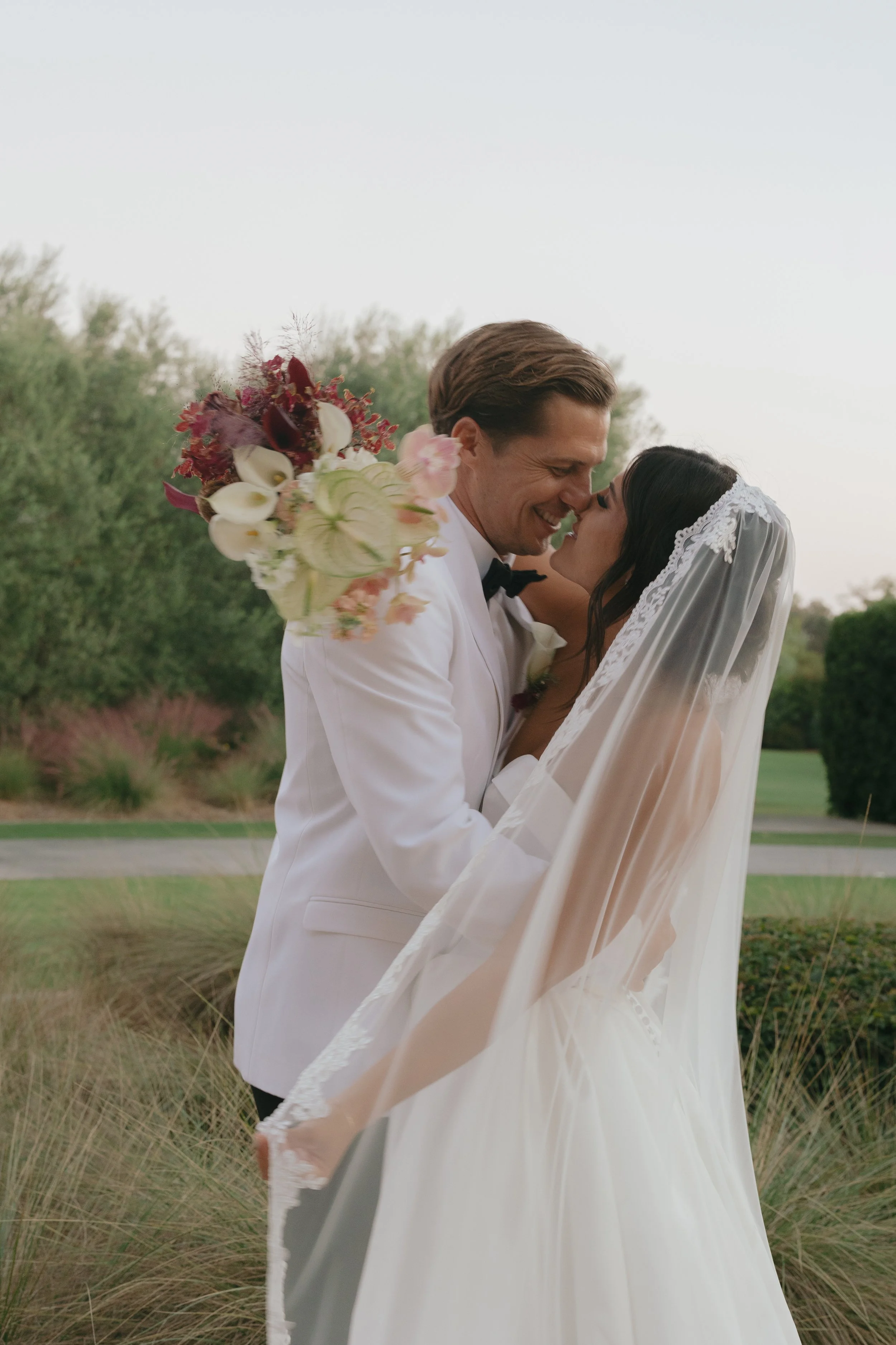 A bride and groom in wedding attire sharing a kiss outdoors, with the groom wearing a white tuxedo and the bride wearing a lace-edged veil and white gown; the groom holds a bouquet of white and purple flowers.