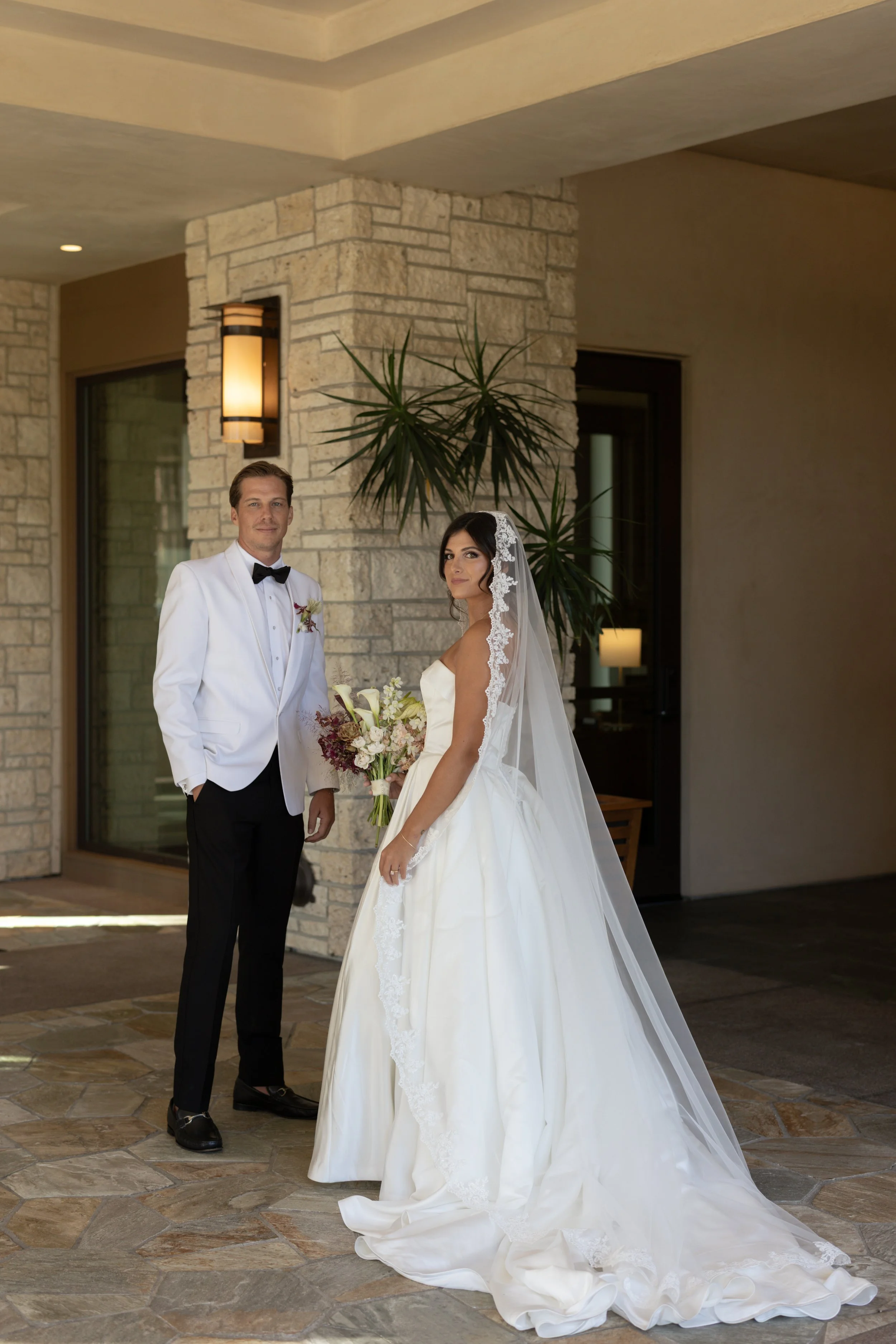 A bride and groom standing inside near a stone wall, with large indoor plants and modern lighting. The bride is in a white wedding dress with a long veil holding a bouquet. The groom is in a black tuxedo with a white jacket and black bow tie.