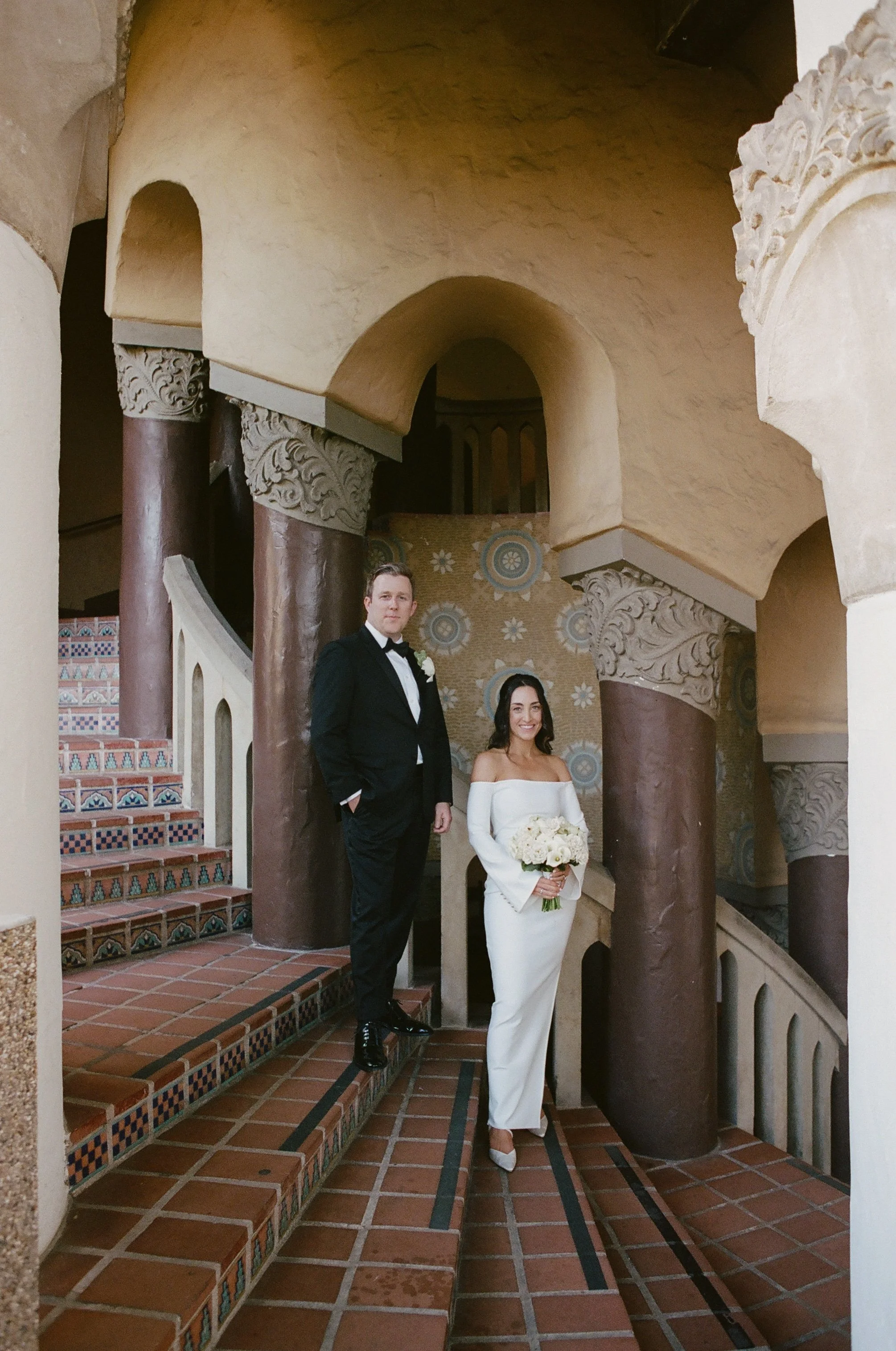 A couple in wedding attire stands on a decorative staircase inside a building with ornate columns and vaulted ceilings; the woman holds a bouquet of white flowers and wears an off-the-shoulder white dress, while the man wears a black tuxedo.