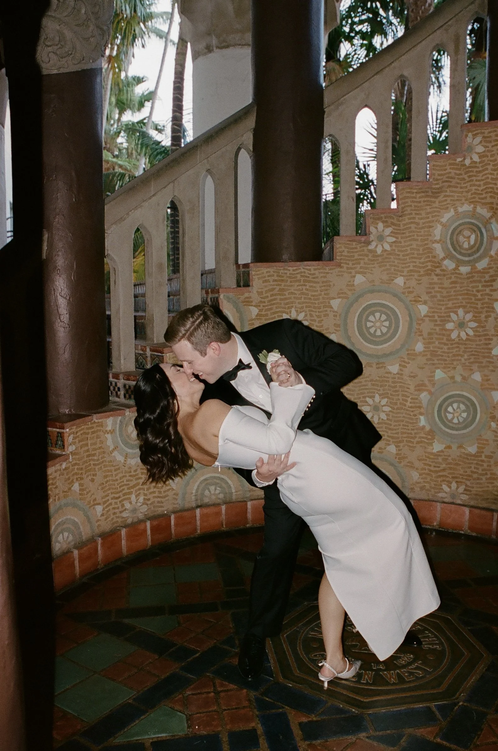 A man in a black tuxedo and a woman in an off-shoulder white dress sharing a dance in a decorative outdoor setting.