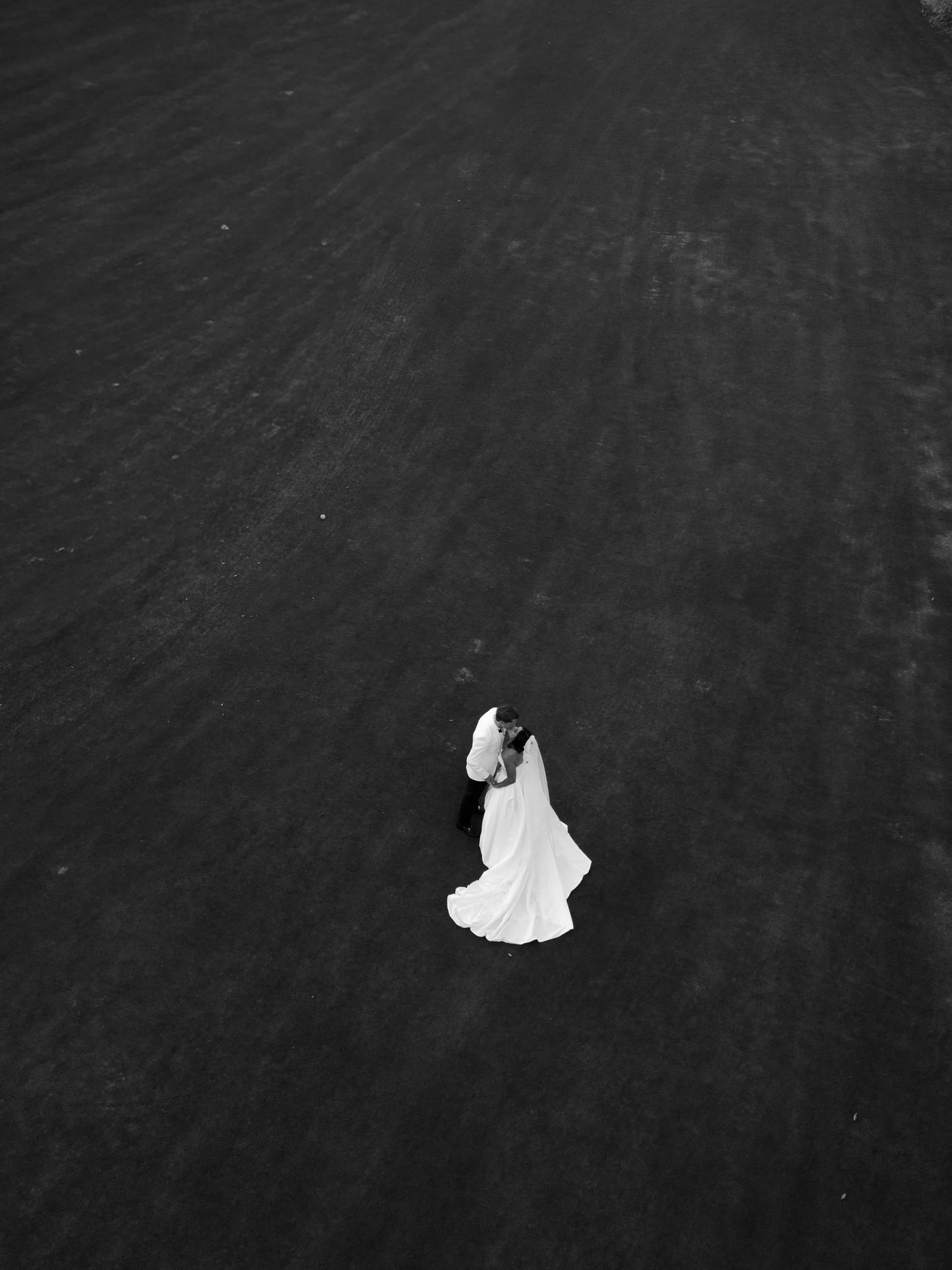 A black-and-white aerial photograph of a bride and groom standing on a large, empty dark surface, sharing a kiss.