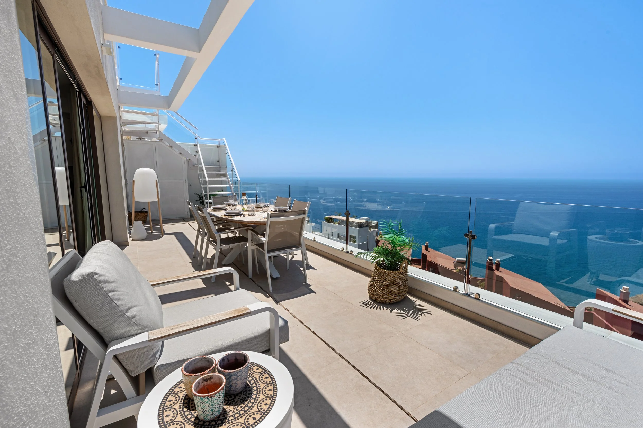 Balcones de apartamento con vista al mar, muebles de exterior, mesa con platos, vasos y plantas decorativas, cielo despejado.