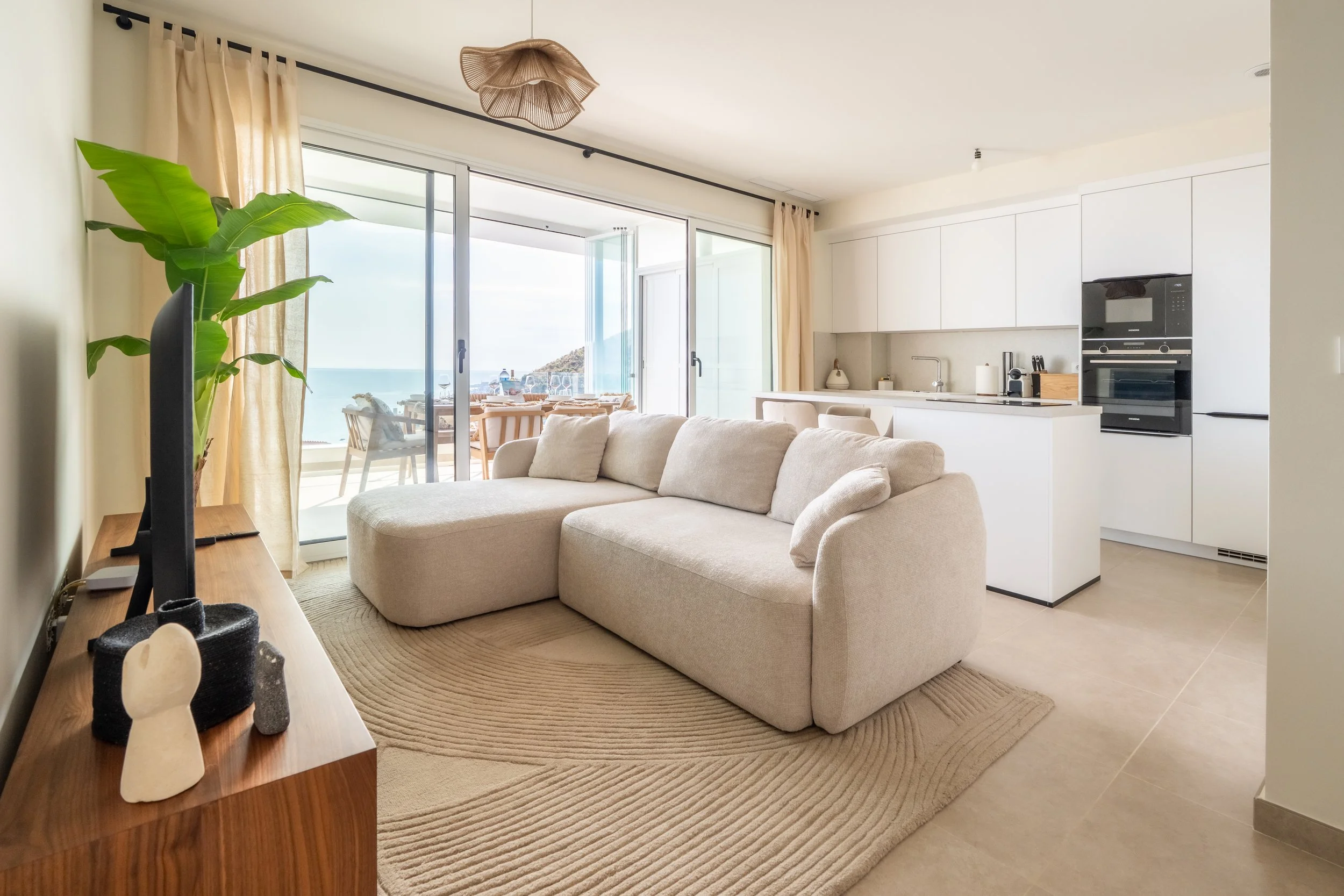 Vista de una sala abierta con sofá beige, visibles ventanas con vista al mar, cocina blanca moderna, y decoración minimalista con plantas y objetos decorativos en la mesa.