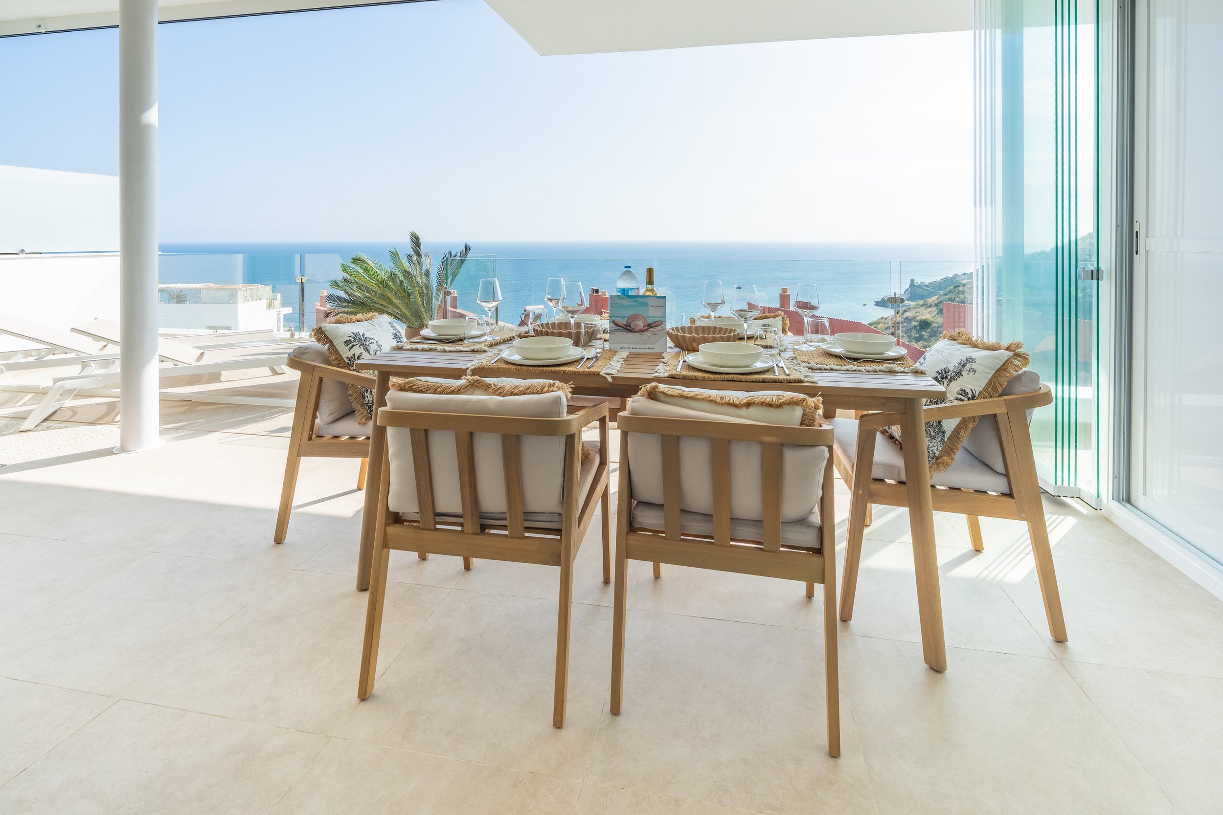 Mesa de comedor en un balcón con vista al mar, con platos, copas y botellas, rodeada de sillas de madera con cojines.
