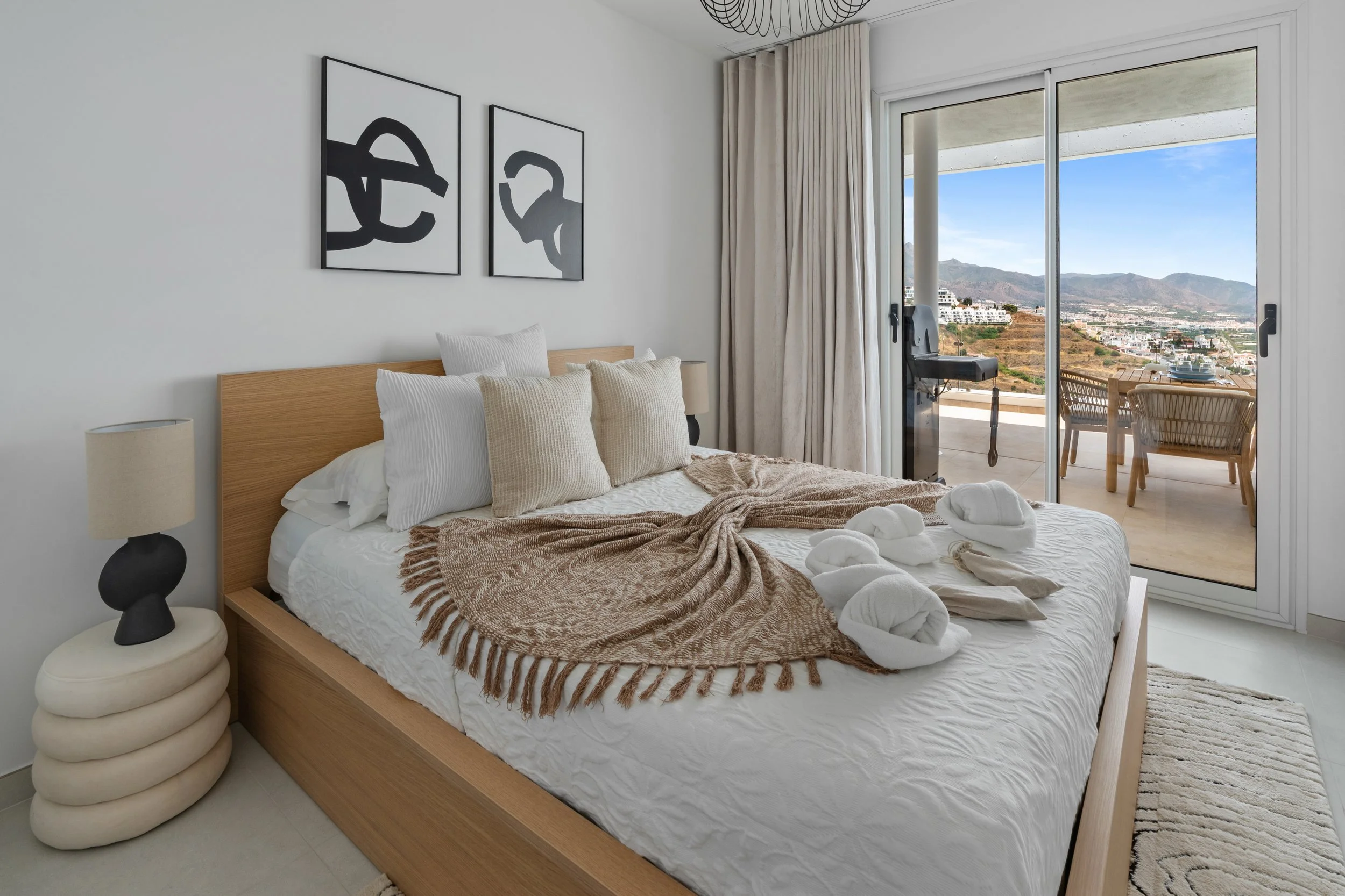 Habitación moderna con cama de madera, almohadas blancas y mullidas, edredón beige, lámpara de mesa negra y beige, cortinas claras, terraza con mesa y sillas, vista a montañas y ciudad, puertas de cristal.