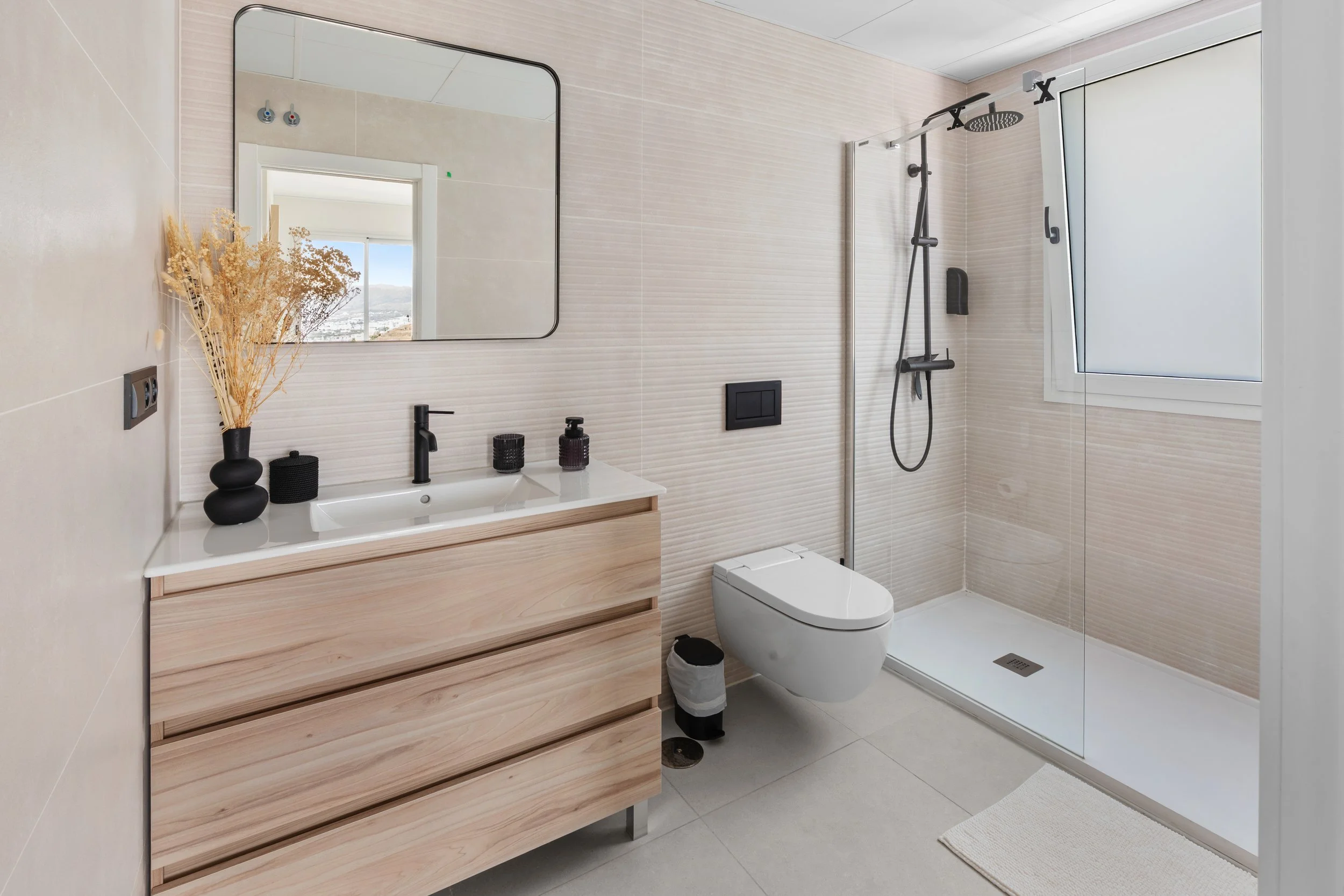 Baño moderno con espejo, lavabo con accesorios, inodoro y ducha, ventanas con vista a la ciudad y decoración sencilla.