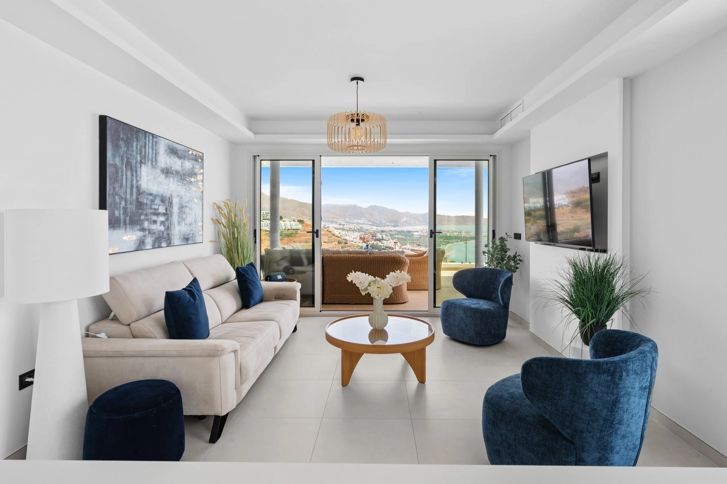 Sala de estar moderna con sillones beige y azules, mesa redonda de madera, plantas decorativas, vista panorámica de montañas y ciudad a través de ventanas grandes, lámpara de techo de diseño de ramas de madera, televisor en la pared, decoración minim