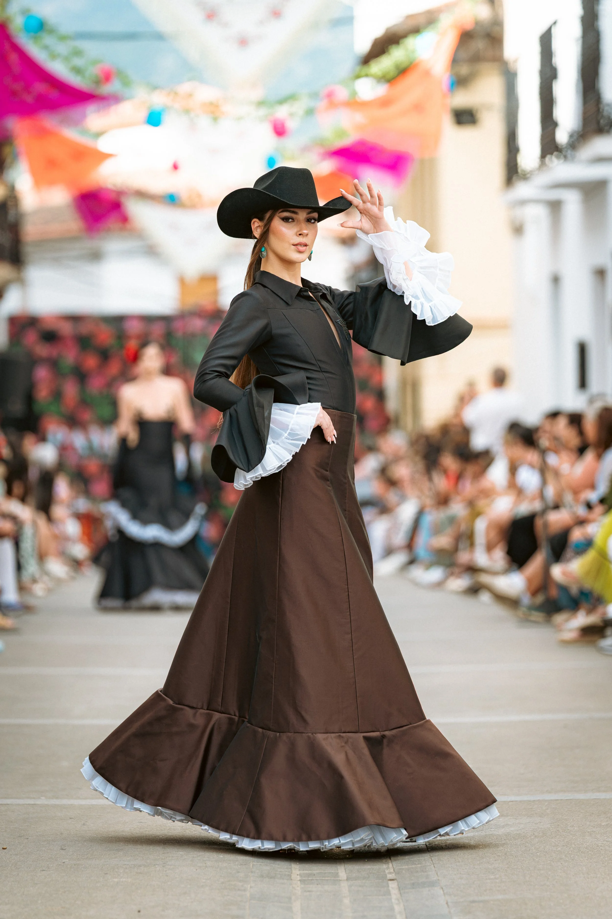 Mujer posando en pasarela con vestido de baile largo de color marrón y blusa blanca con mangas abulladas, lleva sombrero negro y pendientes turquoise en un evento al aire libre. En el fondo, otra modelo vista con un vestido negro y accesorios, con decoraciones de papel de colores colgando sobre la calle.