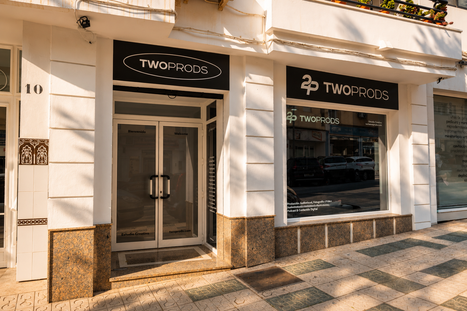 Fachada de un estudio llamada TWOPRODS con dos ventanas grandes y una puerta de vidrio, en una calle soleada, con pavimento decorativo compuesto por baldosas en tonos beige y verdes.