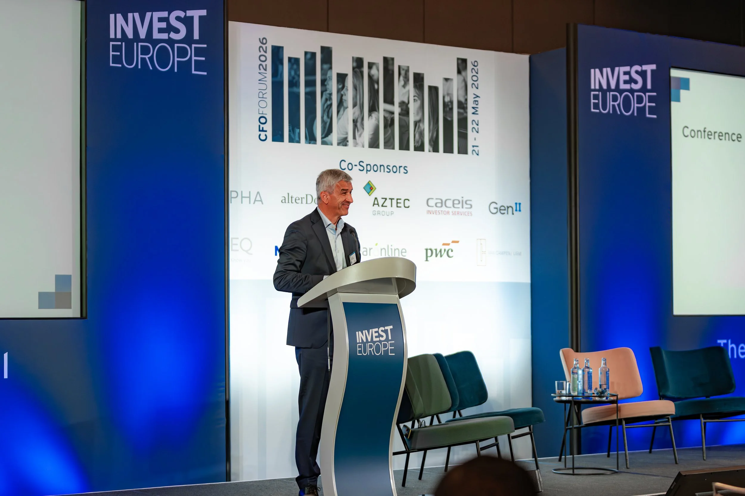 Hombre con chaqueta gris en un podio en una conferencia con cartel que dice 'INVEST EUROPE'. Hay sillas vacías y mesas con botellas de agua y vasos a la derecha.