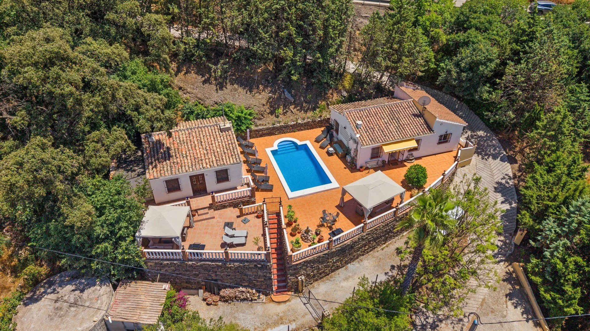 Vista aérea de una casa con piscina, terraza con sombrillas y áreas de descanso, rodeada de árboles y vegetación.