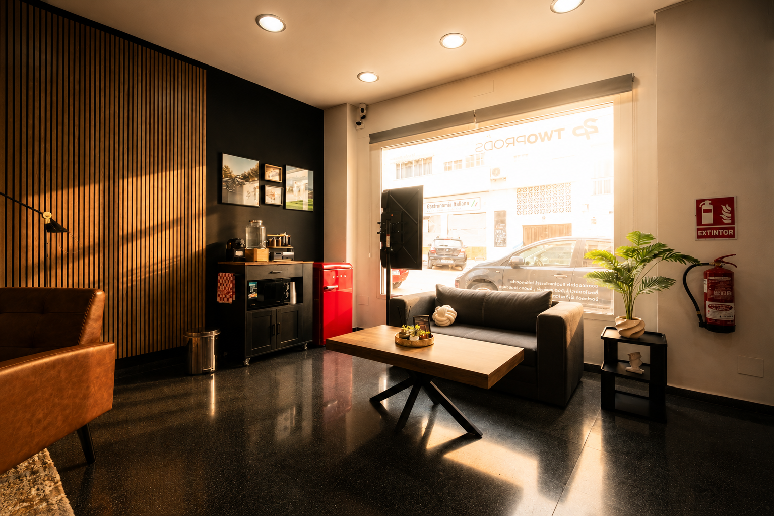 Sala de recepción con sofá negro, mesa de madera, plantas y decoración moderna, con ventanas que muestran autos y edificios afuera.