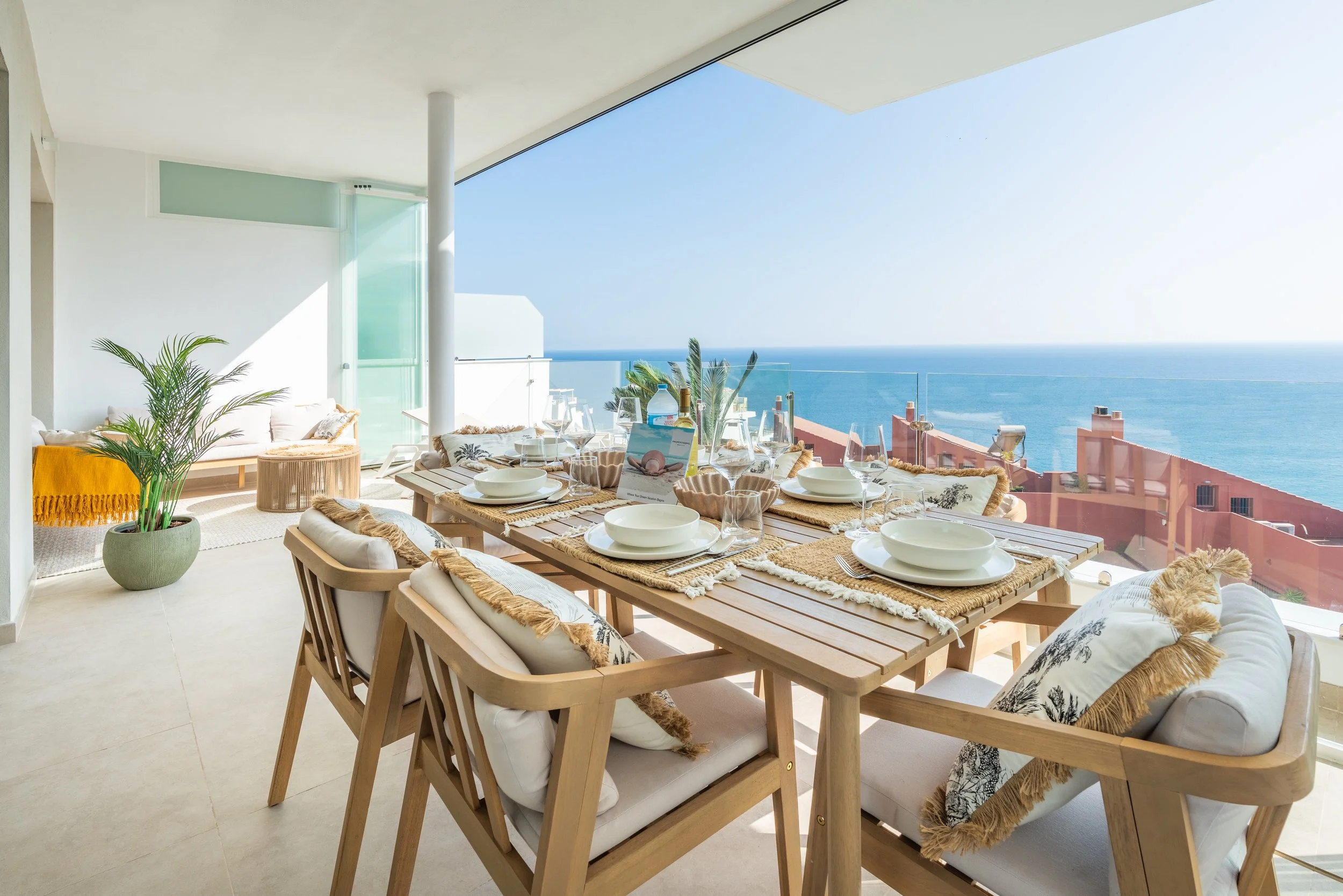 Comedor en balcón con vista al mar, mesas con vajilla, plantas y muebles de madera, ambiente luminoso y moderno.