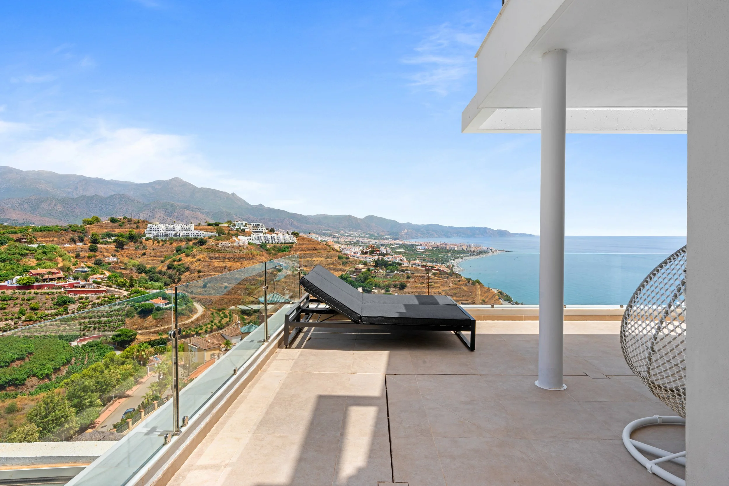 Vista de una terraza con vista al mar, con una silla de mimbre blanca y una tumbona negra, bajo un techo blanco con columna, y un paisaje de montañas y casas en contextura mediterránea.
