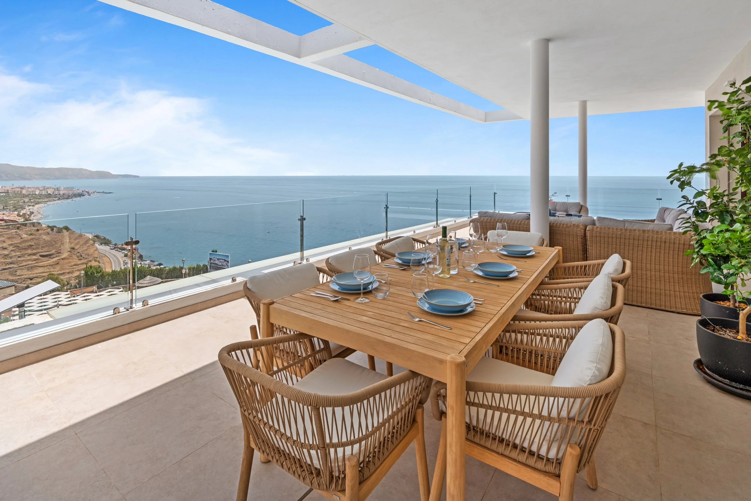 Comedor al aire libre con vista al mar, mesa de madera con platos y copas, sillas de mimbre, plantas y estructura moderna.