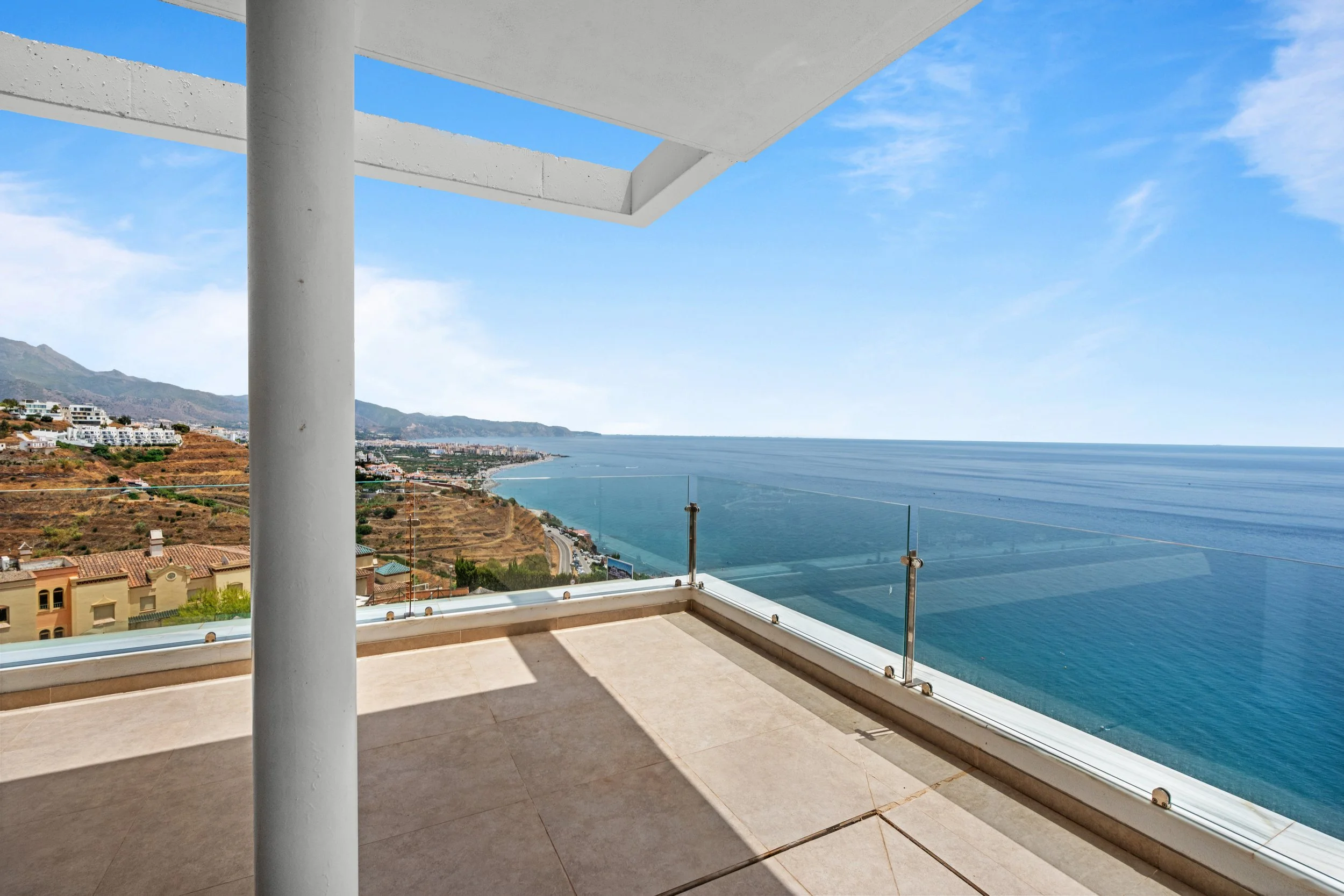 Vista de un balcón con barandal de cristal, con el océano y la costa en el horizonte, y colinas con casas en la distancia.