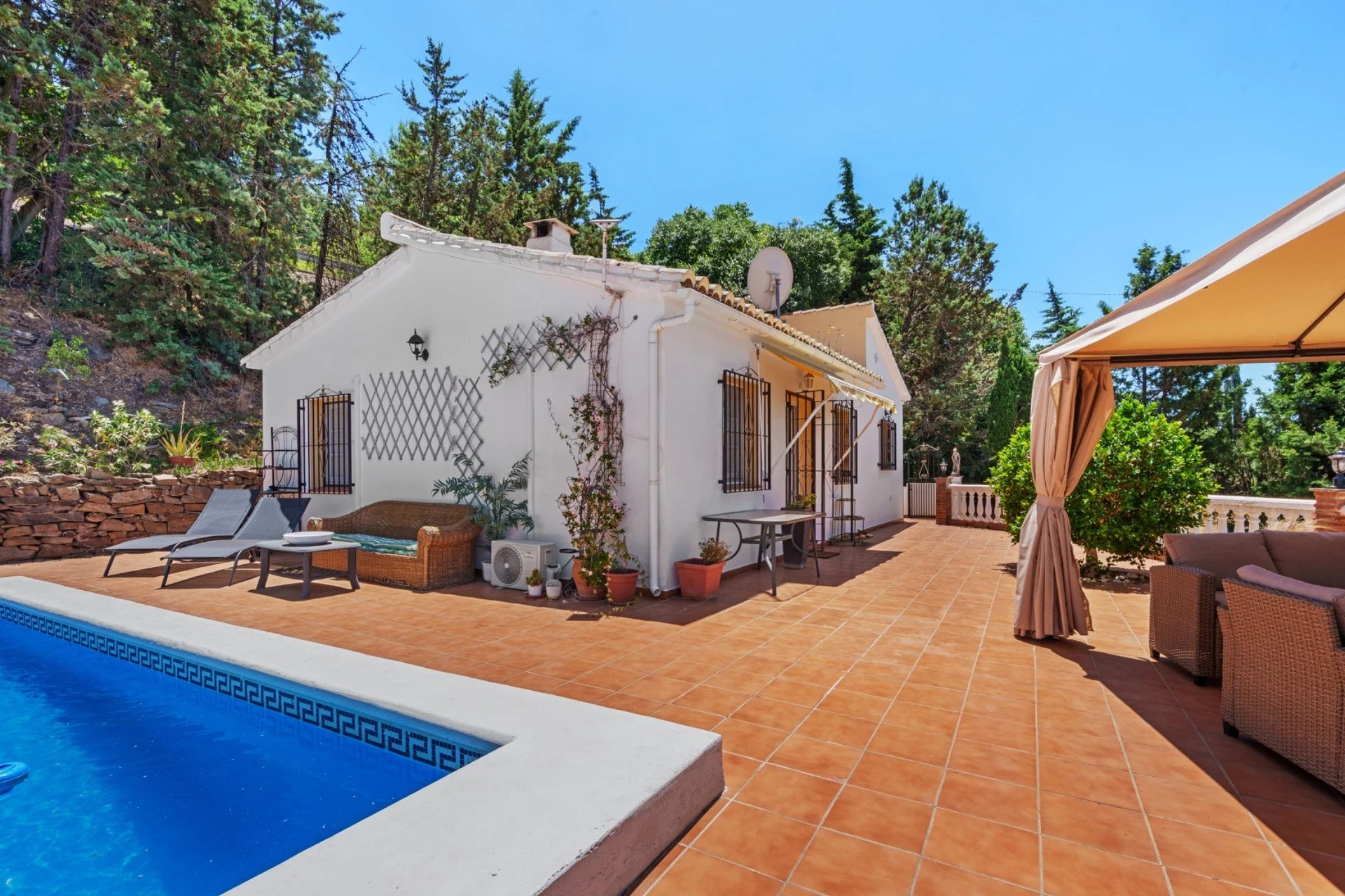 Casa blanca con ventanas con rejas, terraza con sillones y sombrilla, piscina y árboles verdes bajo un cielo azul
