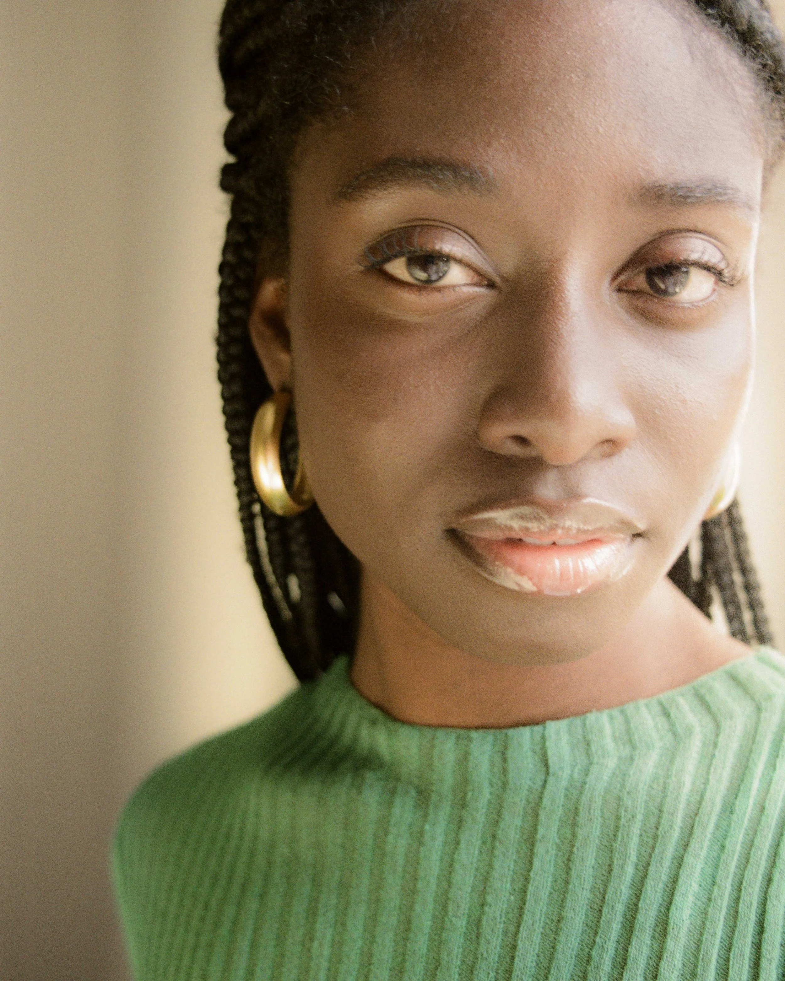 Close-up portrait of a young Black woman with braided hair, wearing gold hoop earrings and a green ribbed sweater, looking slightly to the side.