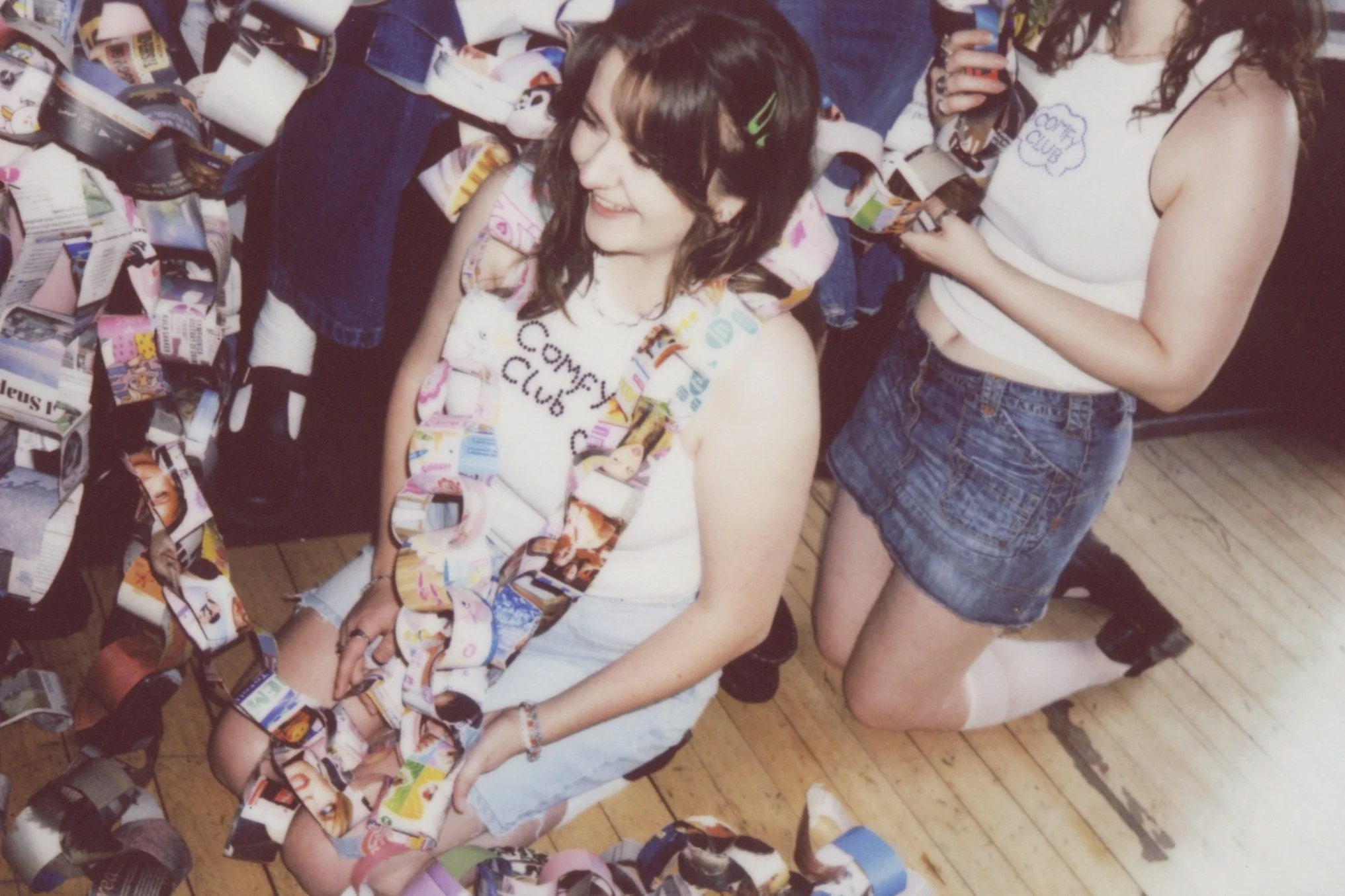 A girl kneeling on the wooden flooe with a smile, wearing a white tank top with 'Comfy Club' written on it, surrounded by strips of colourful magazines or paper. 
