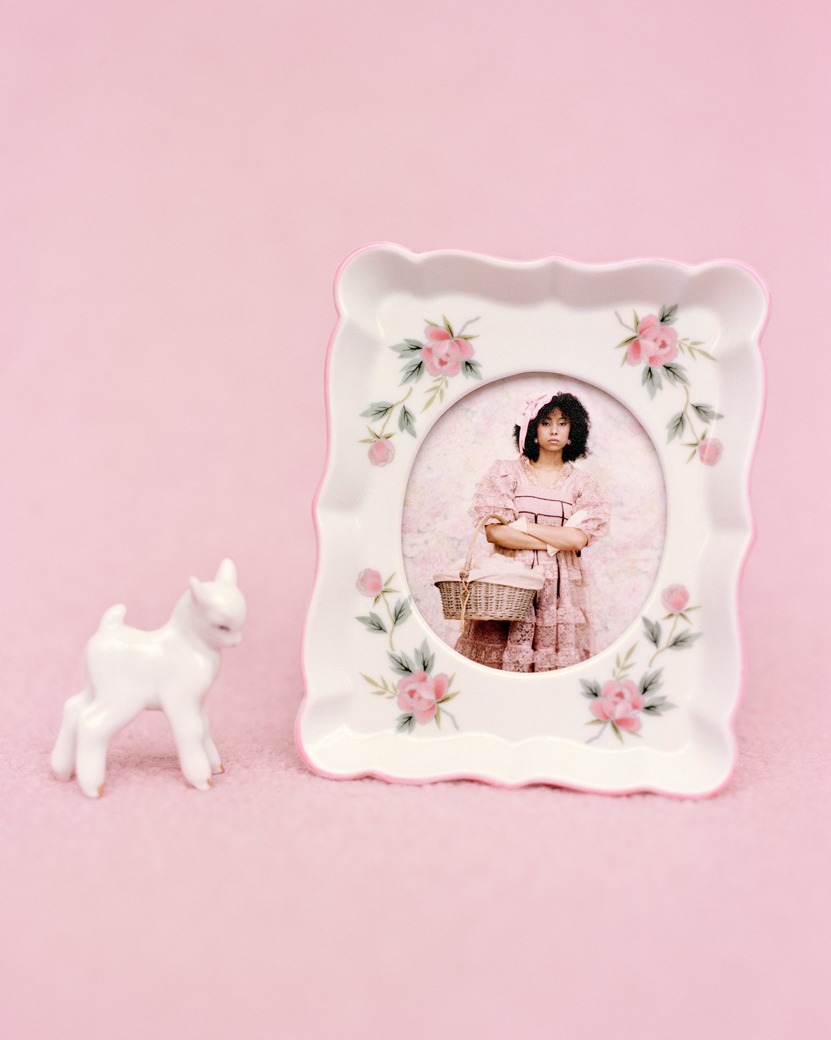 A decorative picture frame with pink floral patterns, containing a photo of a young woman in pink clothing holding a basket, against a pink background. A small white ceramic lamb figure is placed next to the frame.