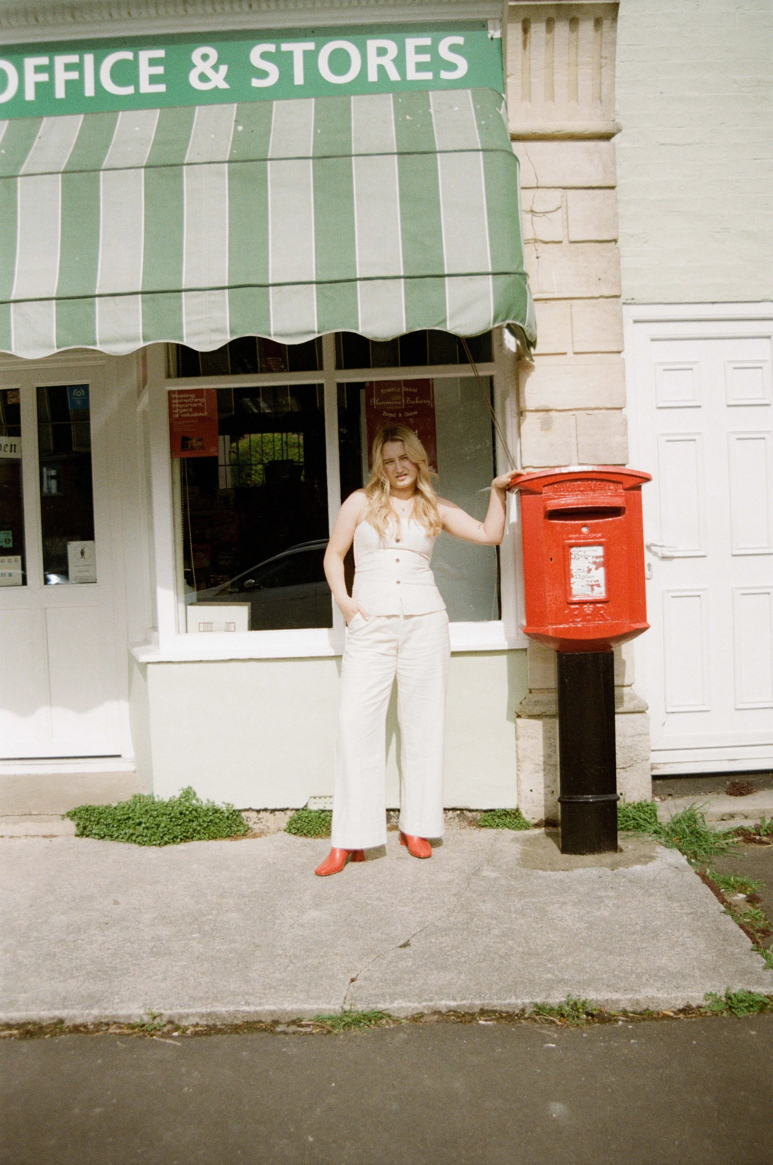 A woman standing outside a building with a green awning that reads 'Office & Stores,' next to a red mailbox.