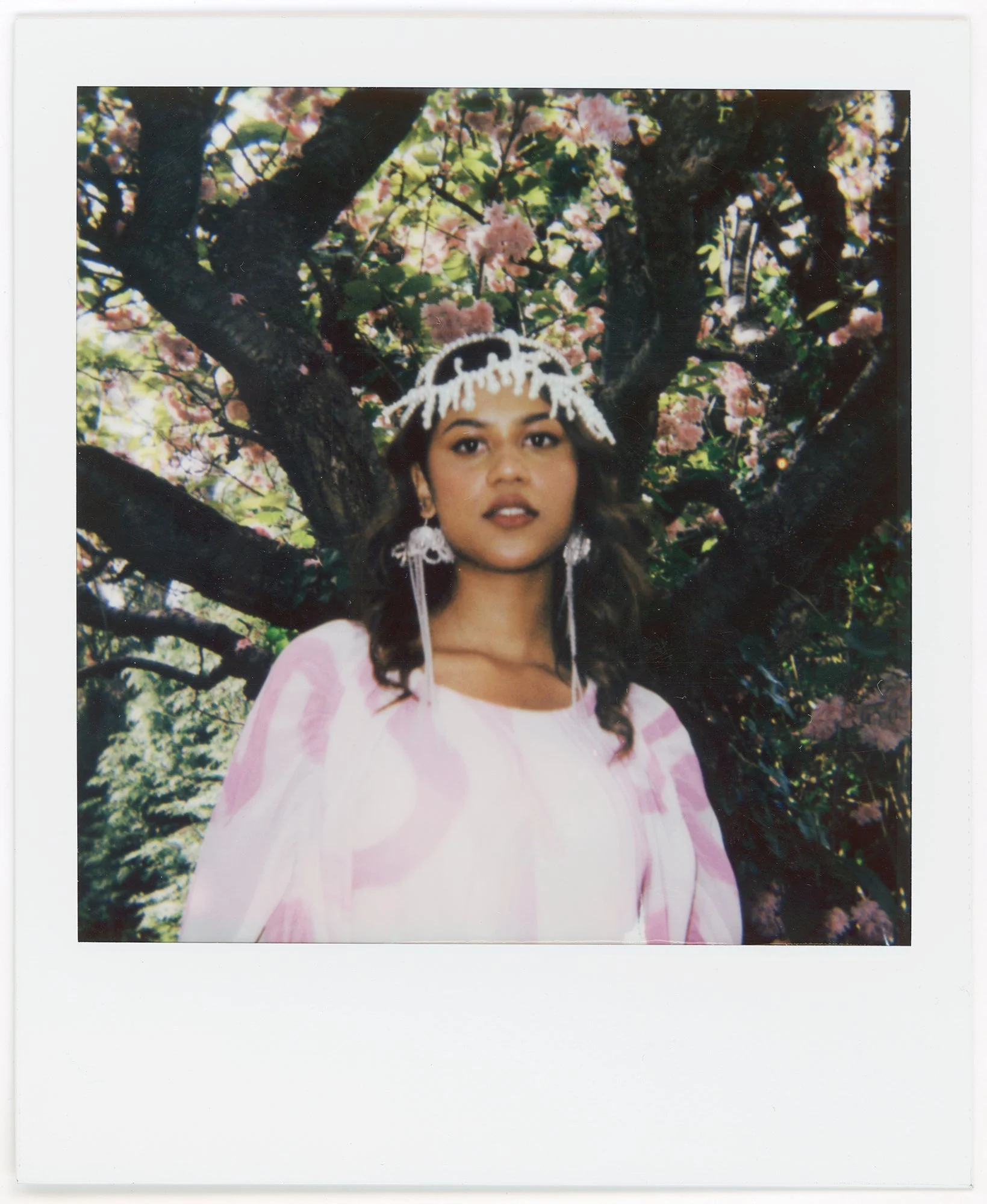 A young woman with dark hair and earrings stands in front of a blossoming pink-flowered tree, wearing a white dress with pink patterns and a white decorative headpiece.