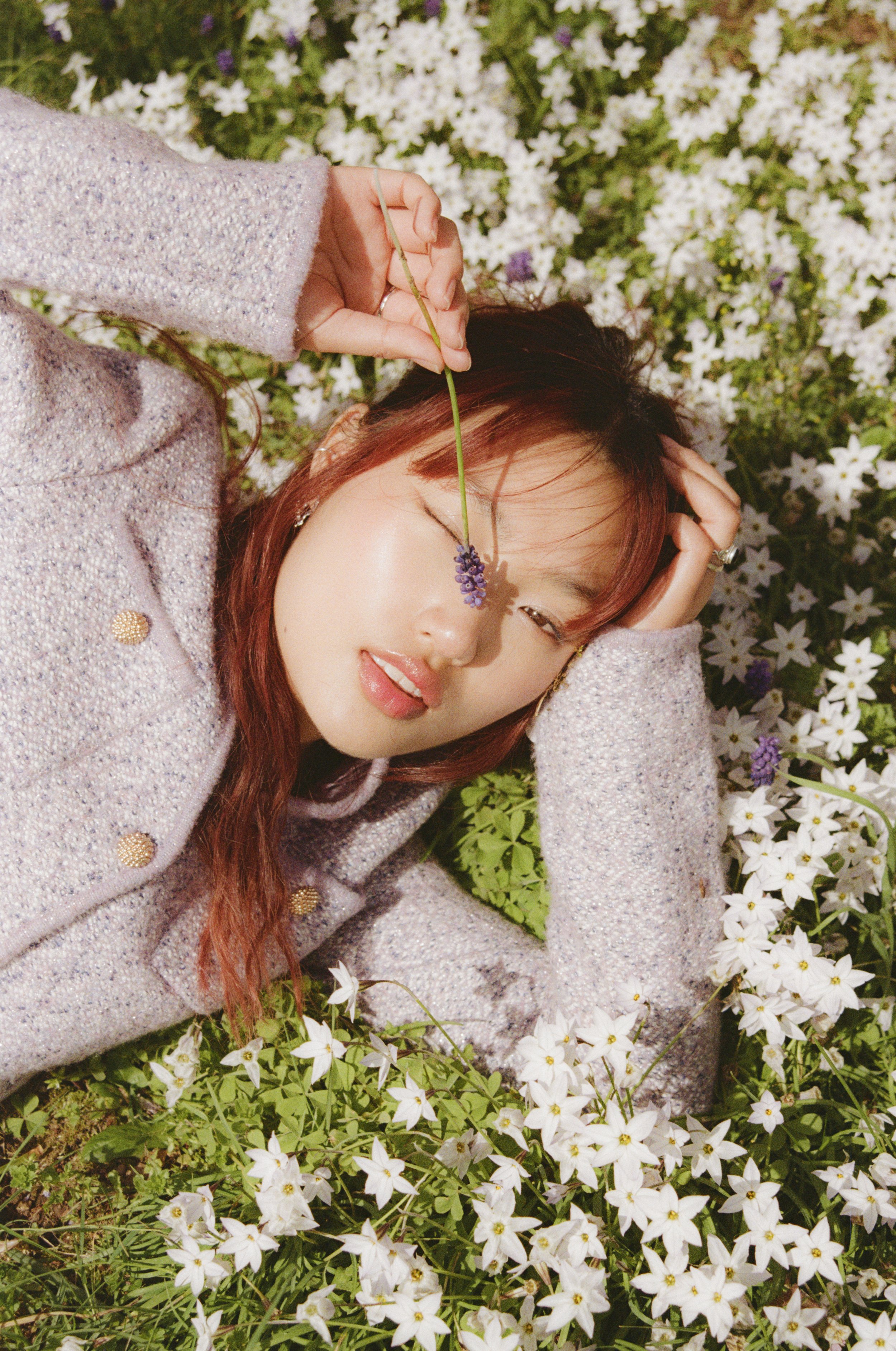 A woman with reddish hair lies among white flowers, holding a purple flower stem over her face, winking and looking relaxed.
