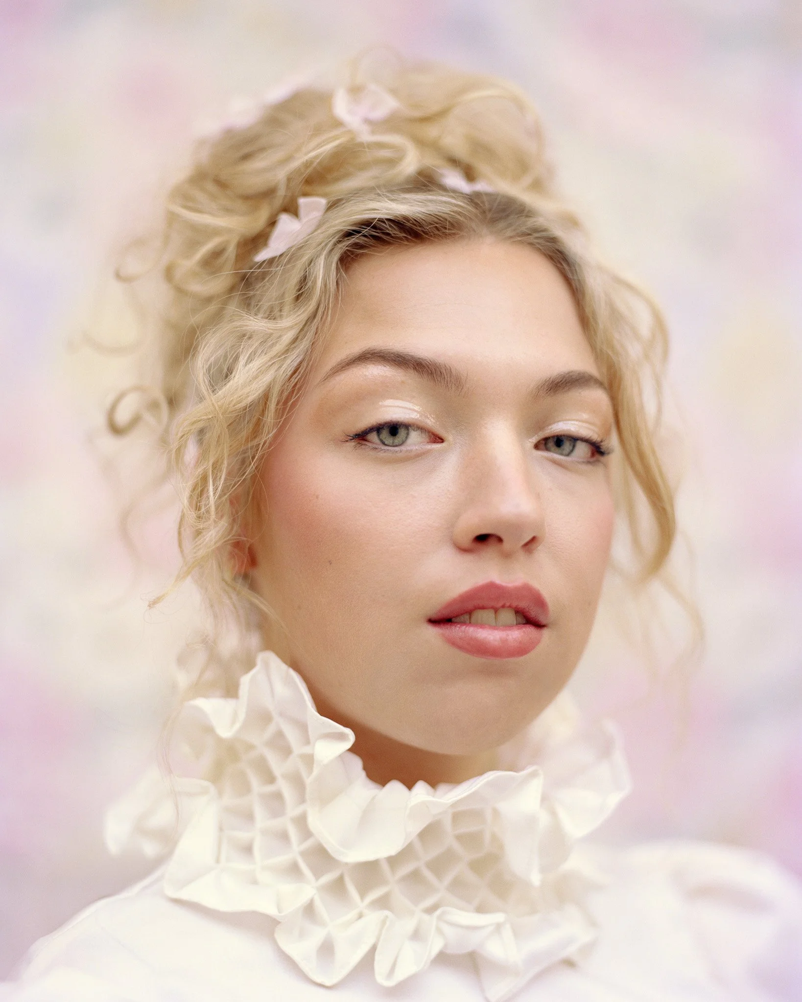 Close-up of a woman with curly blonde hair styled with pink ribbons, wearing a ruffled white collar, and makeup with soft pink lipstick and eyeshadow.