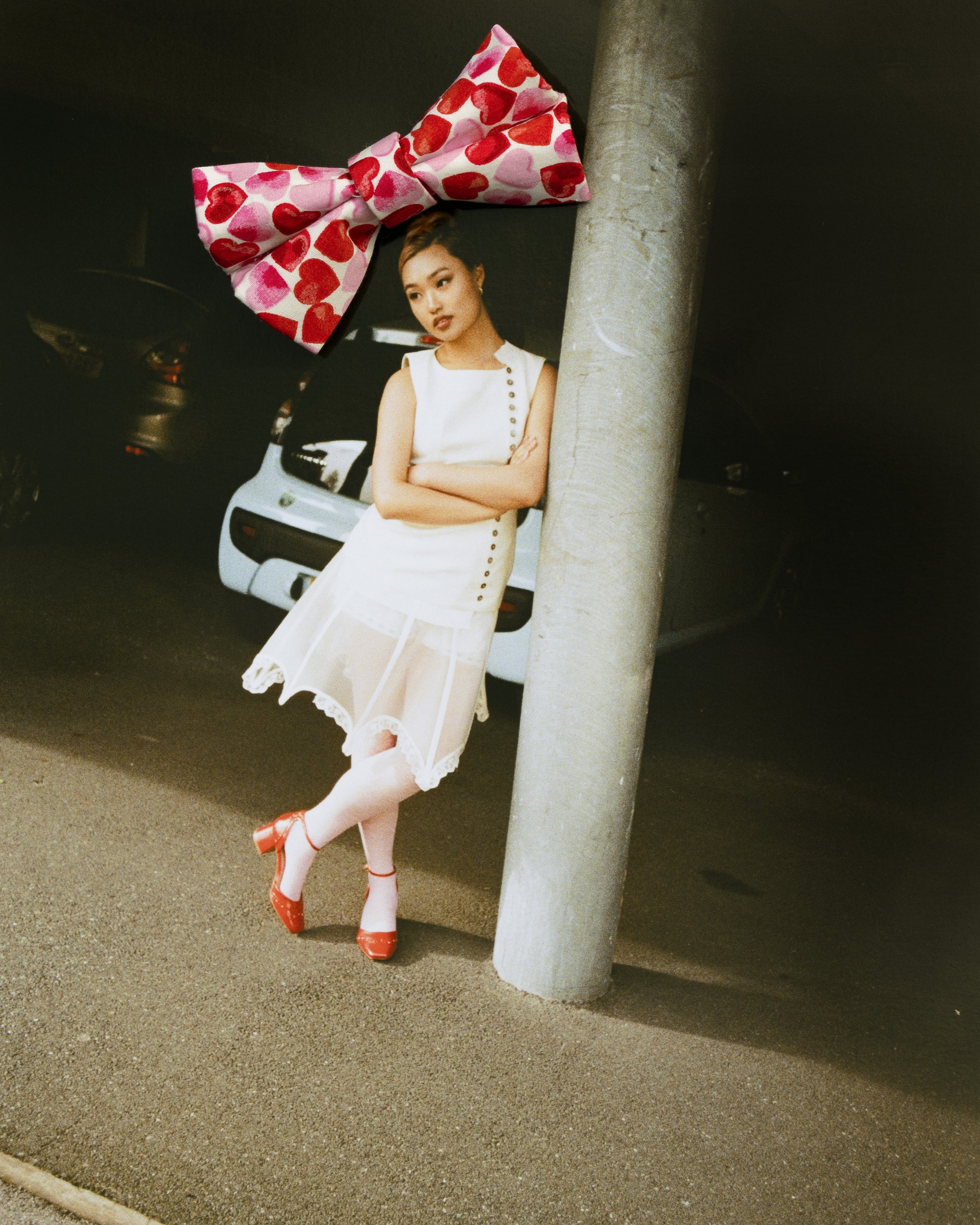 A woman in a white dress with a sheer skirt, red high heels, and a large pink bow with red hearts on her head, leaning against a concrete pole in a parking garage.