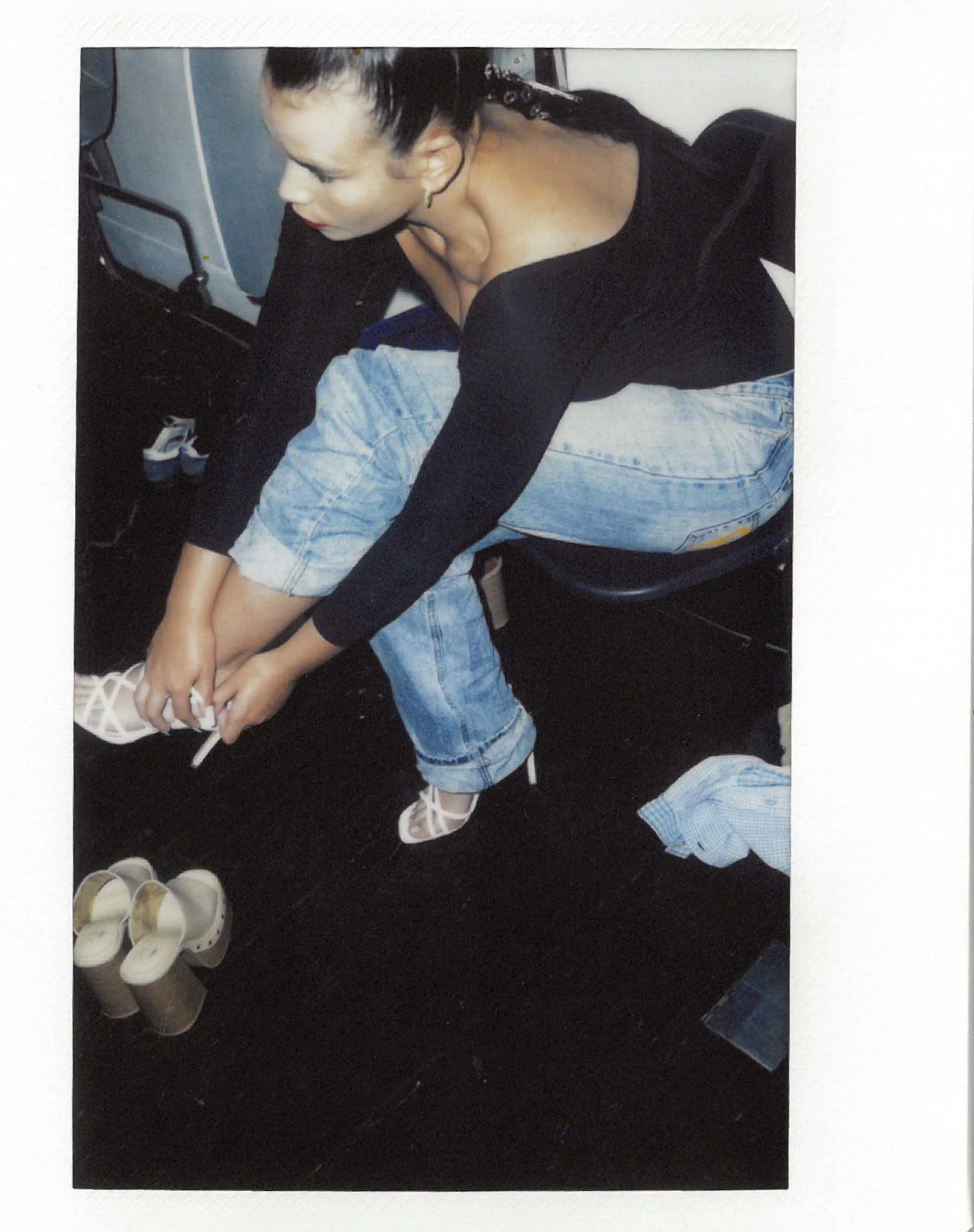 A woman sitting and tying her shoelaces, wearing a black top and ripped jeans, with several pairs of shoes on the floor near her.