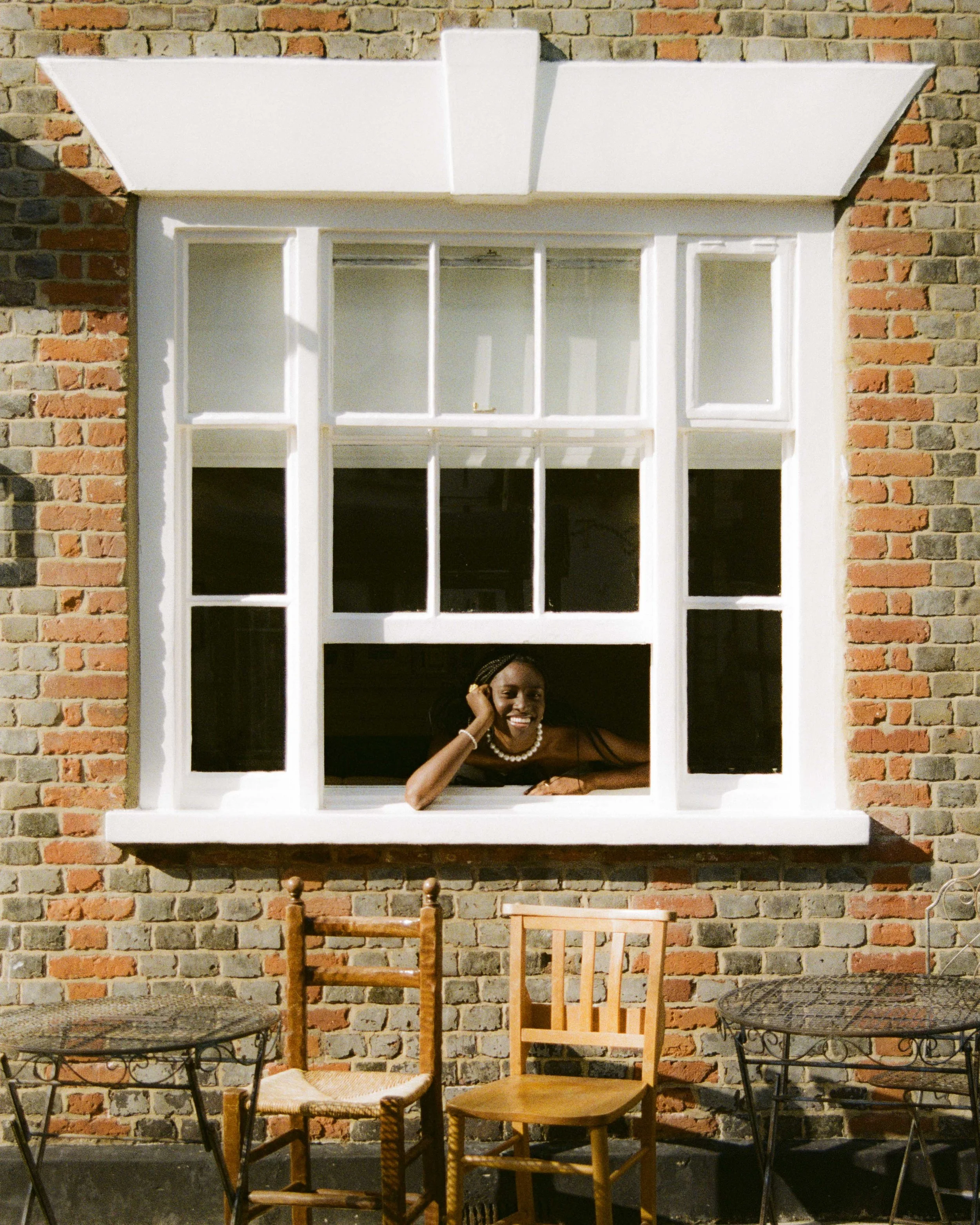 A woman smiling and leaning her head on her hand through a large open window in a brick wall, with outdoor tables and chairs in front.