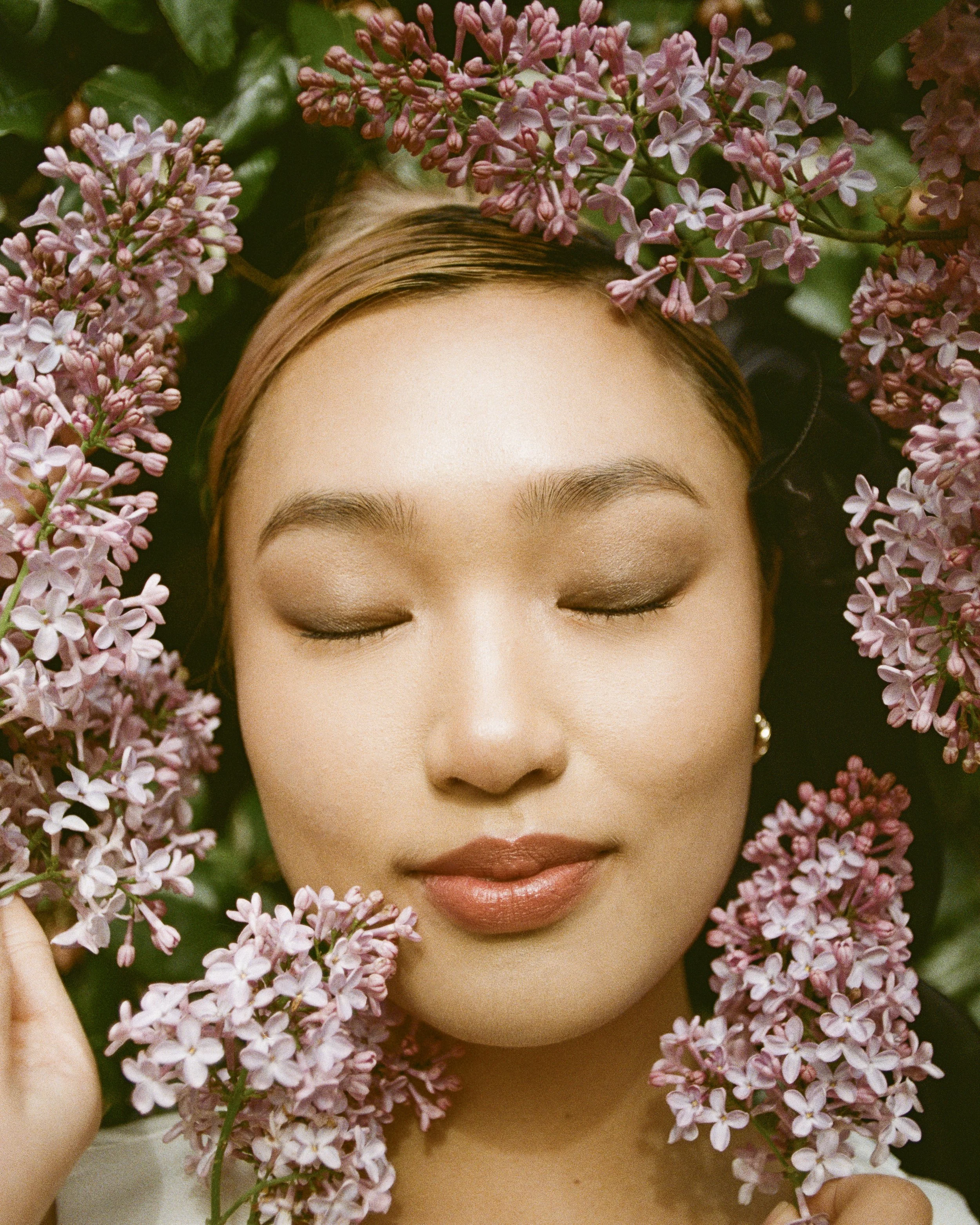 A woman with closed eyes and makeup, surrounded by pink lilac flowers.