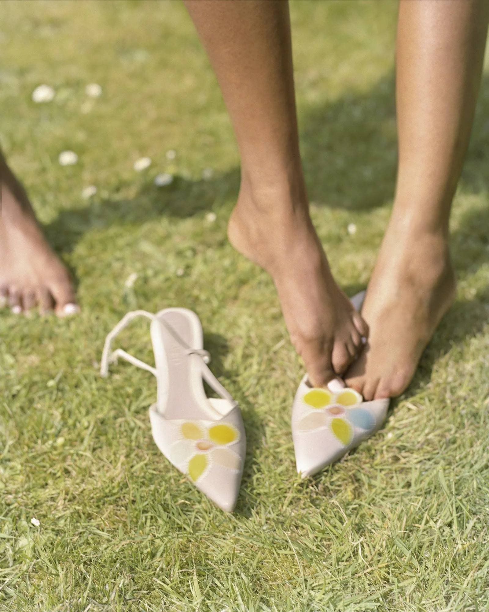 Two woman with light brown skin are standing barefoot on grass, both trying to fit into the same kitten heels which are pink with a yellow and pink floral pattern. Another shoe is on the ground nearby.