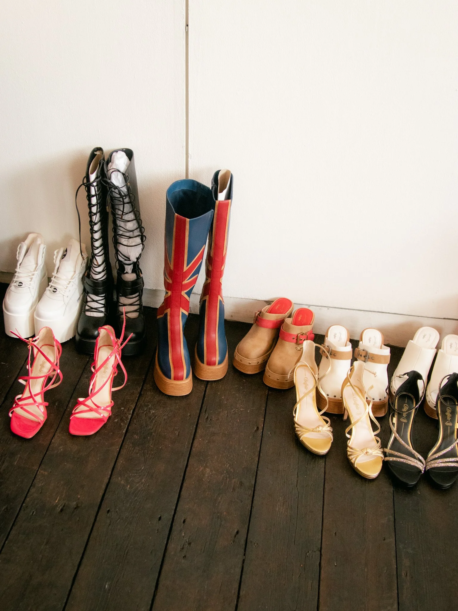 A row of various footwear including boots, high heels, clog heels, gold strappy sandals, and black sandals with rhinestones, all lined up on a dark wooden floor against a white wall.