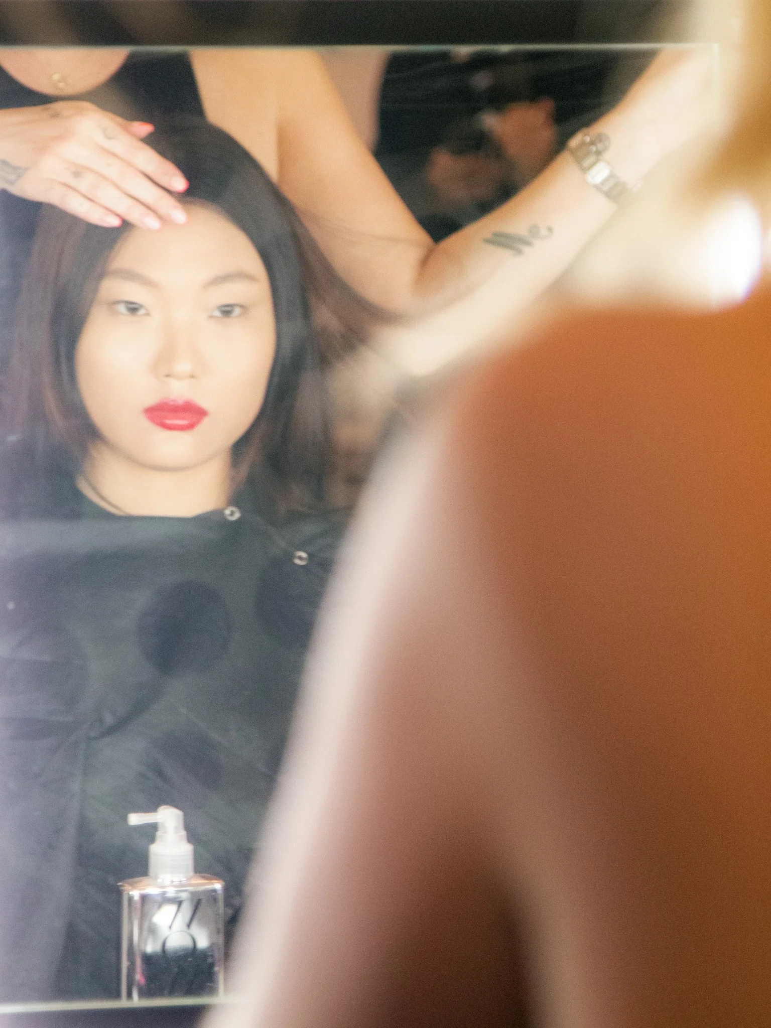 A woman with shoulder-length dark hair and red lipstick is sitting in front of a mirror while a stylist straightens her hair. The stylist's arm and hand are visible, with a tattoo on her wrist and a bracelet.