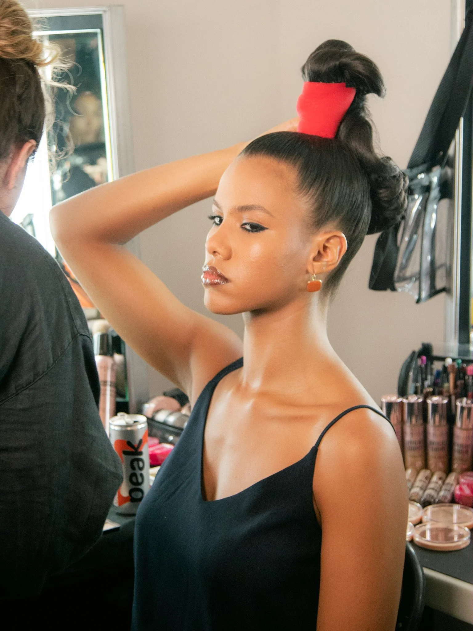 A woman with a high, intricately styled updo with a red accessory, wearing a black spaghetti strap top, getting her hair and makeup done in a studio or backstage setting.