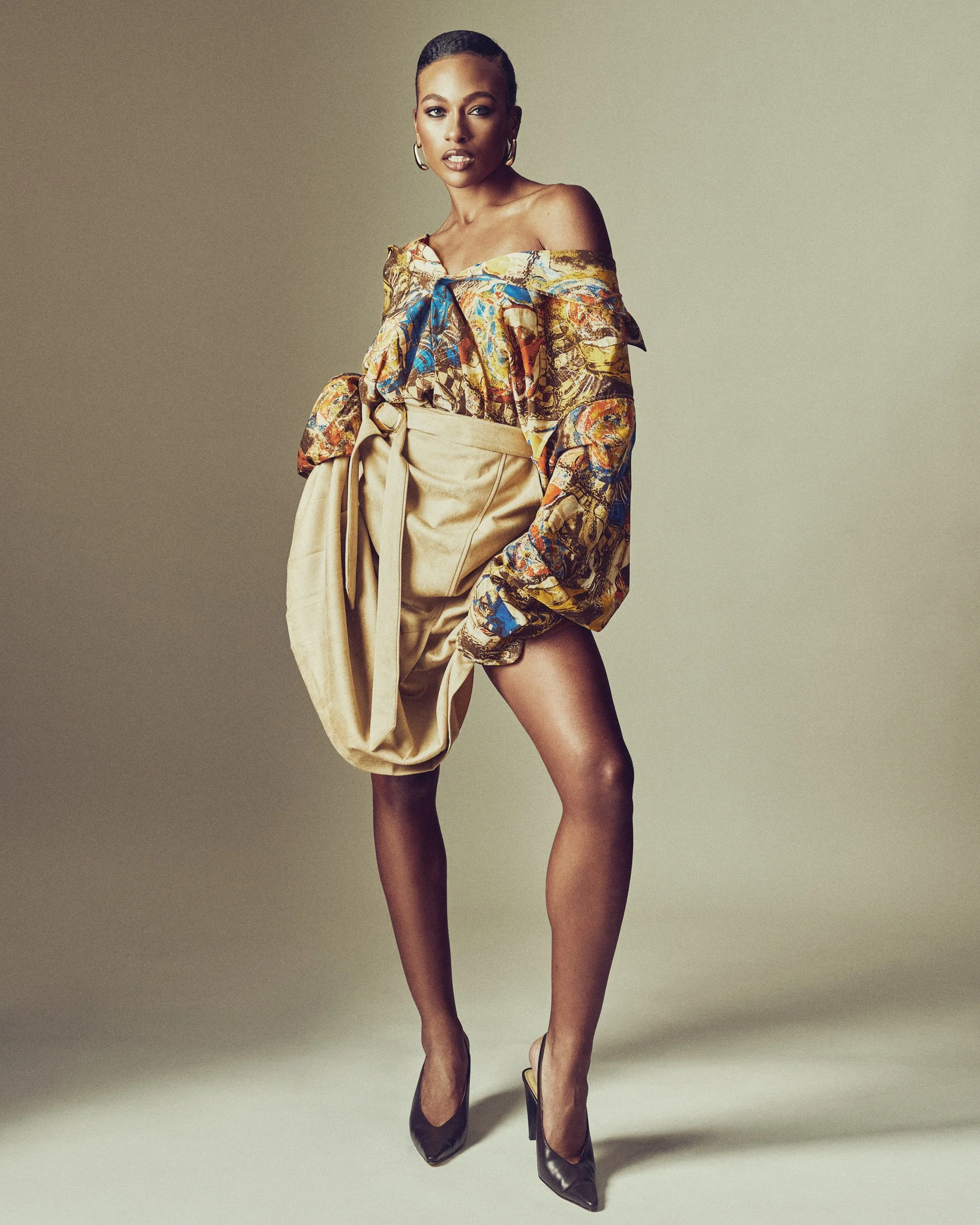 A woman wearing a colorful off-the-shoulder top with voluminous sleeves and a beige skirt with a bow, high heels, standing against a neutral background.