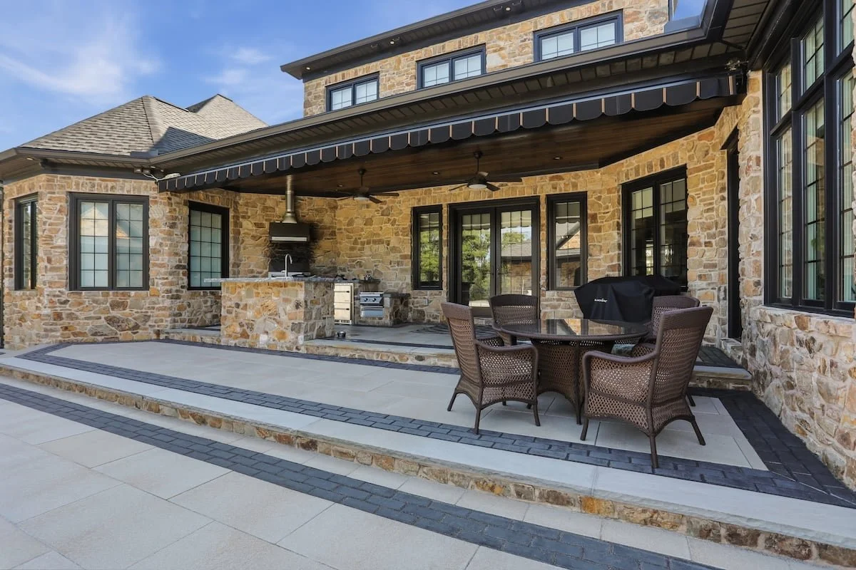 Covered stone patio with a wicker dining set beside an outdoor kitchen island — Weaver's Landscape Company