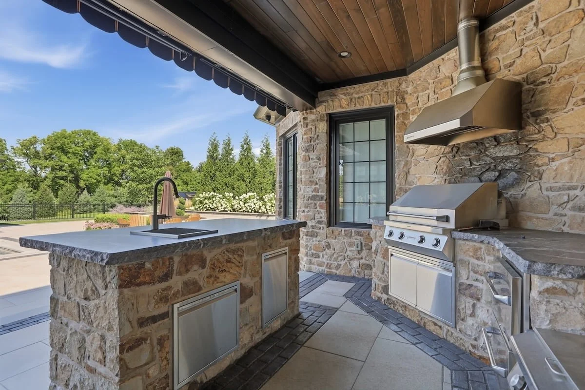 Covered stone patio with a wicker dining set beside an outdoor kitchen island — Weaver's Landscape Company