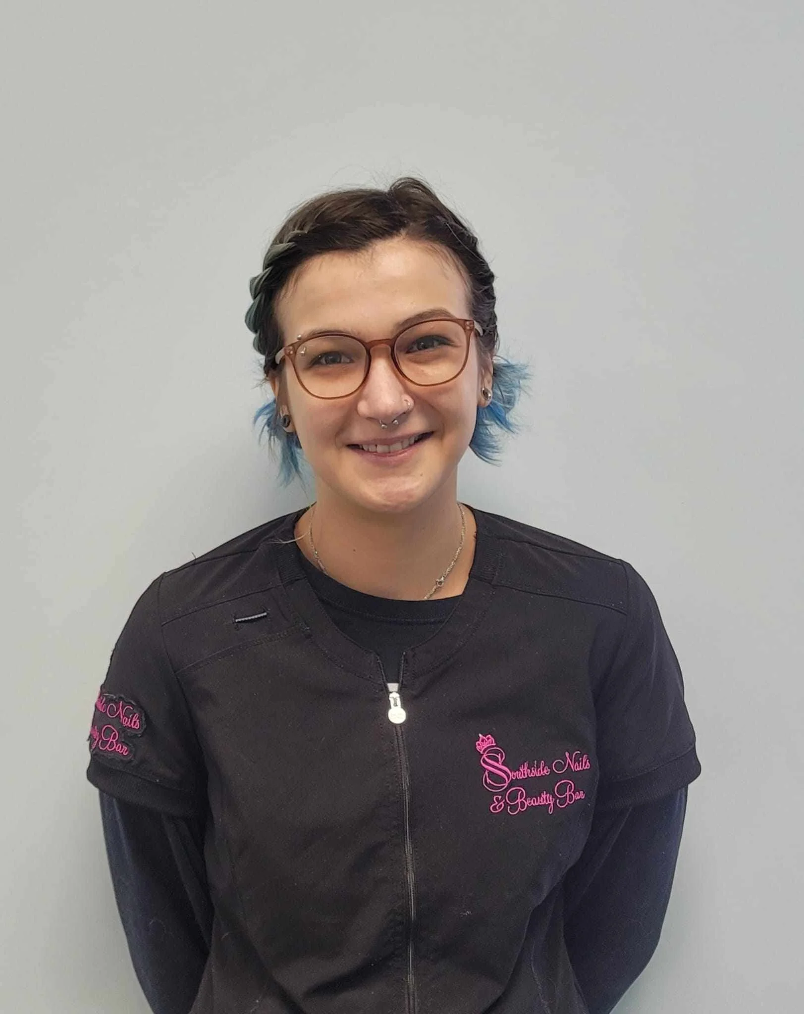 Portrait of a young woman with short dark hair, blue tips, glasses, and a septum piercing, smiling at the camera, wearing a black uniform with pink embroidery that says 'Southside Nails & Beauty Bar'.