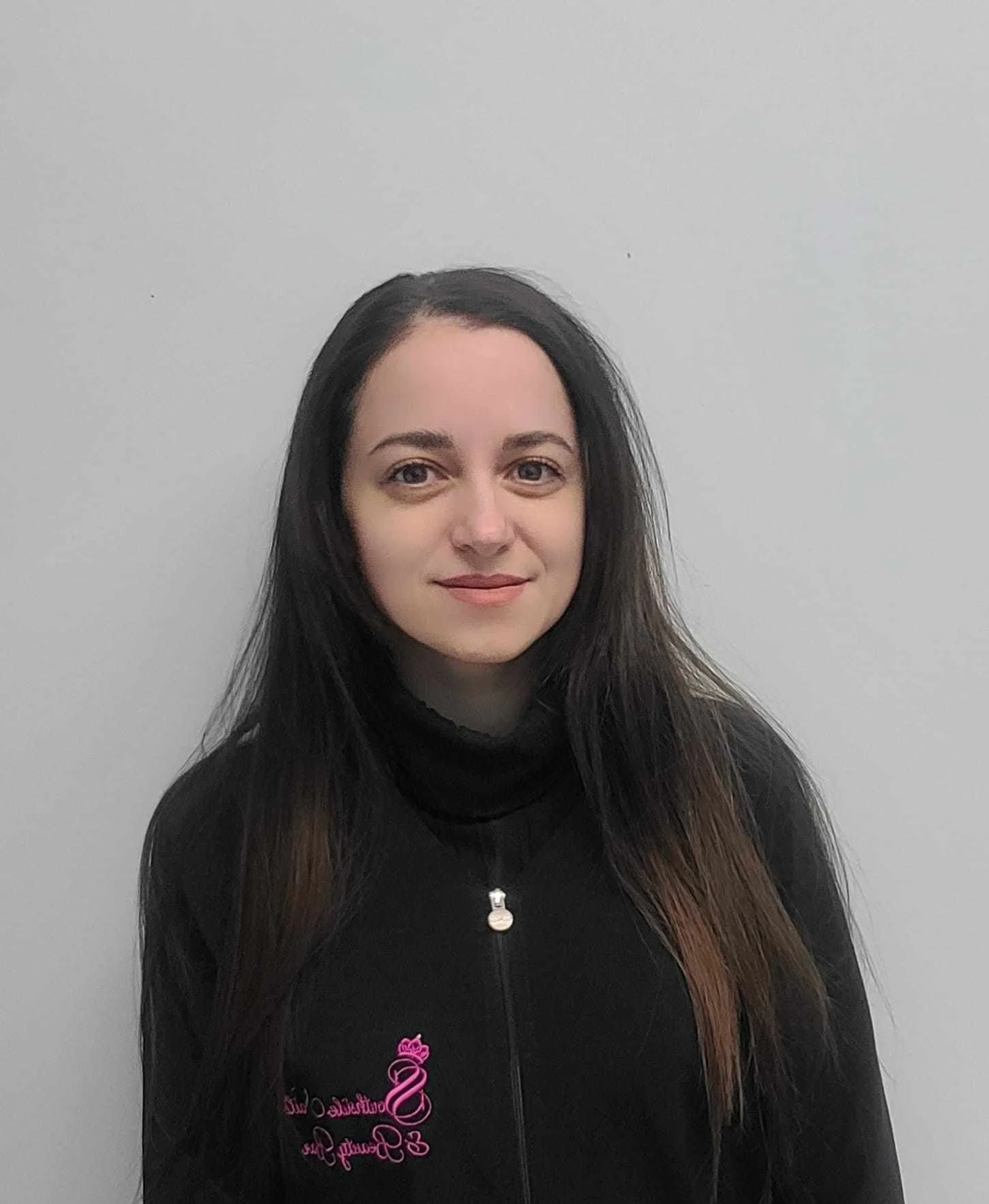 A young woman with long dark hair standing against a plain light gray wall, wearing a black zip-up jacket with pink embroidery.