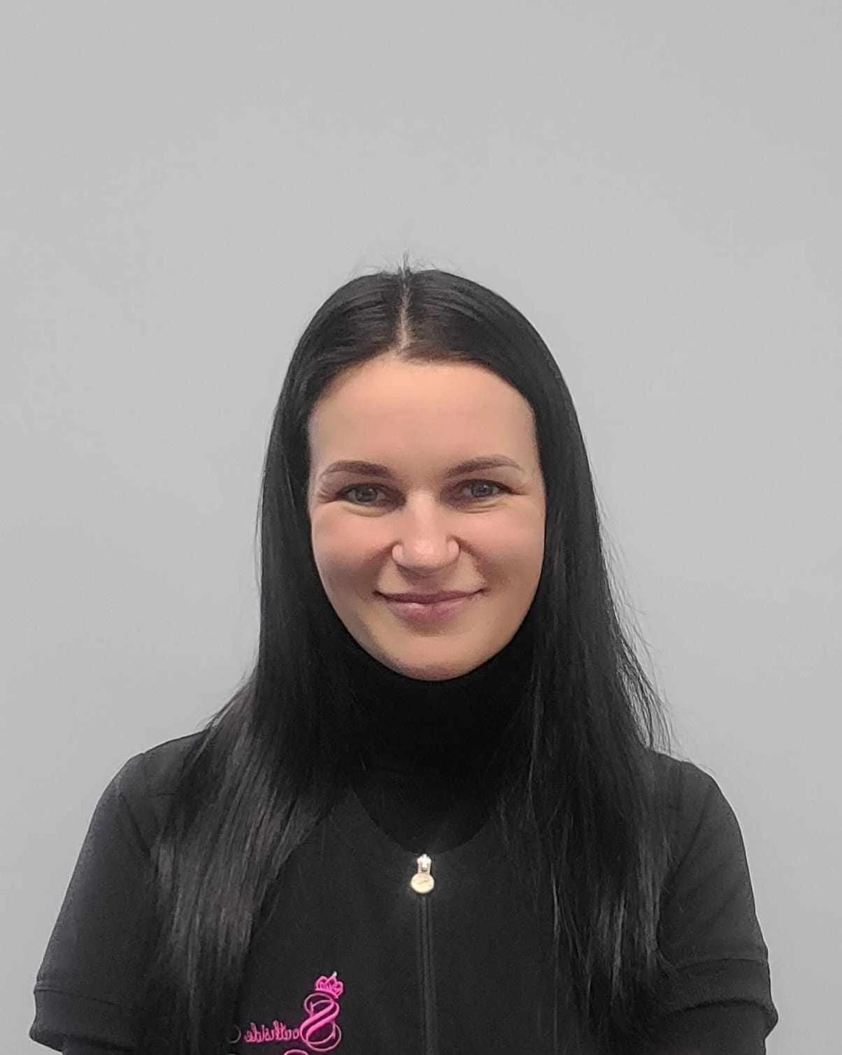 Portrait of a smiling woman with long black hair, wearing a black zip-up jacket with pink embroidery, standing against a plain light gray background.