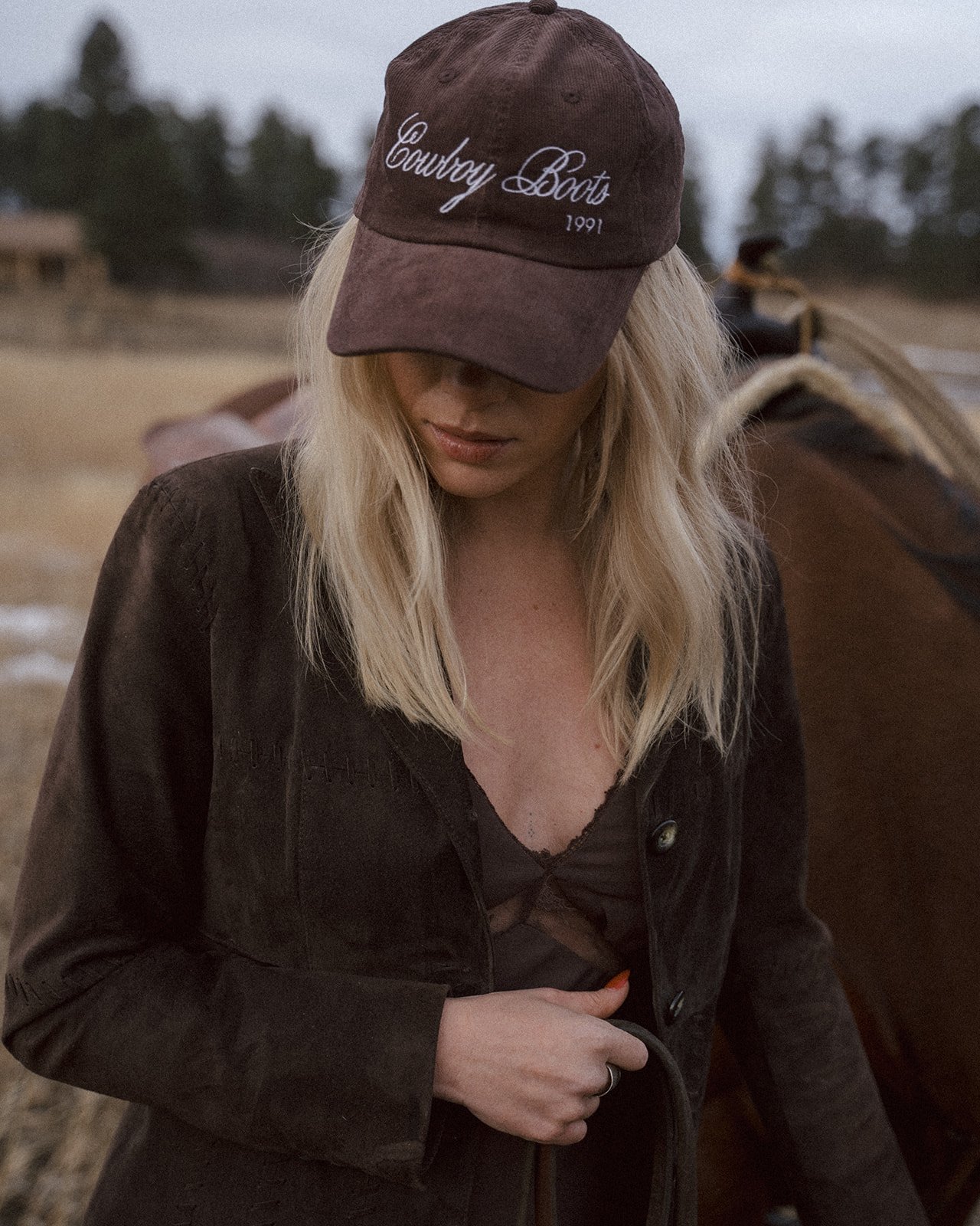 Person wearing a brown cap with 'Cowboy Boots 1991' embroidered, brown jacket, standing near a horse in an outdoor setting with trees in the background.