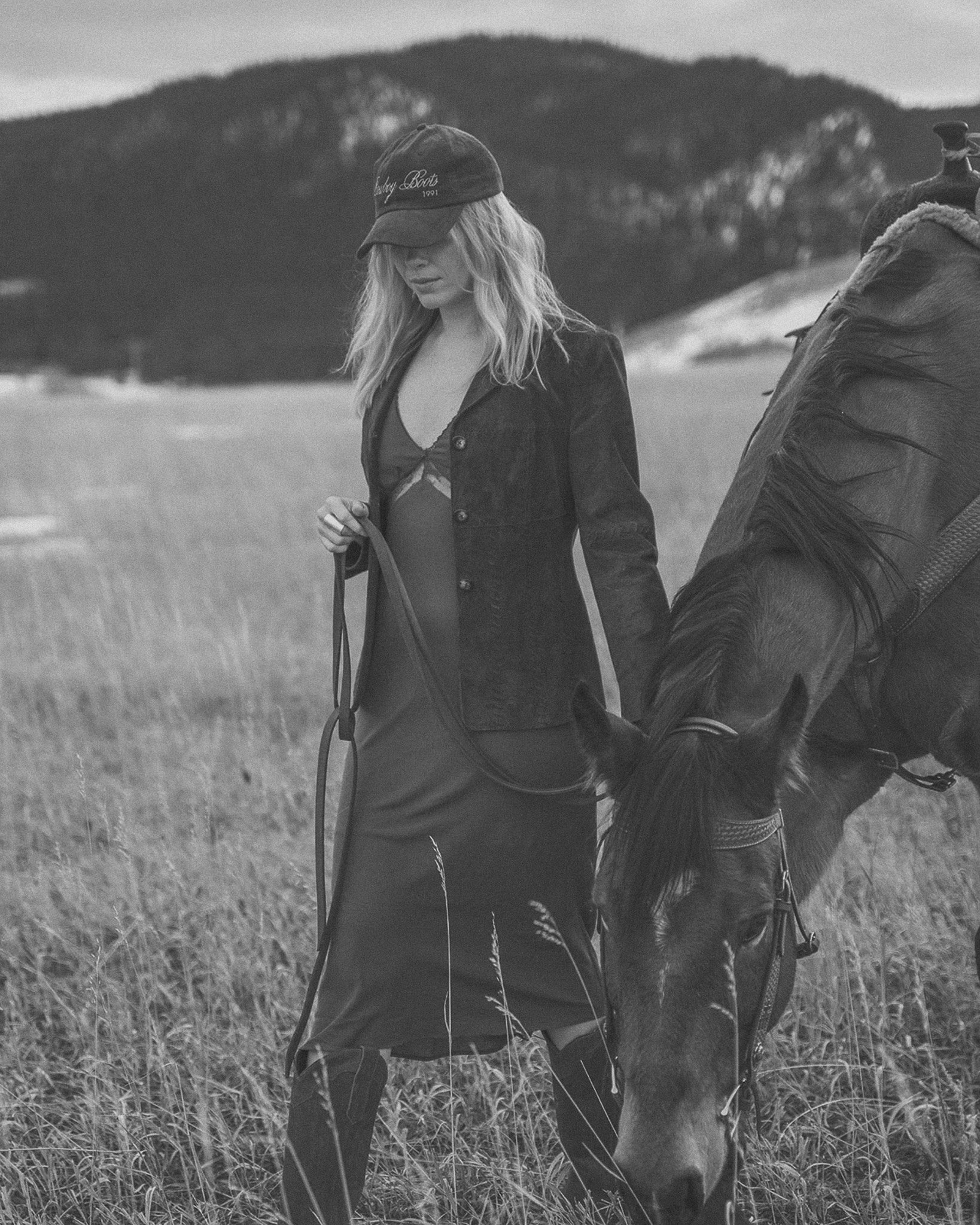 Black and white image of a woman in a field holding the reins of a horse. She is wearing a long coat, a dress, and a cap. A hilly landscape is in the background.