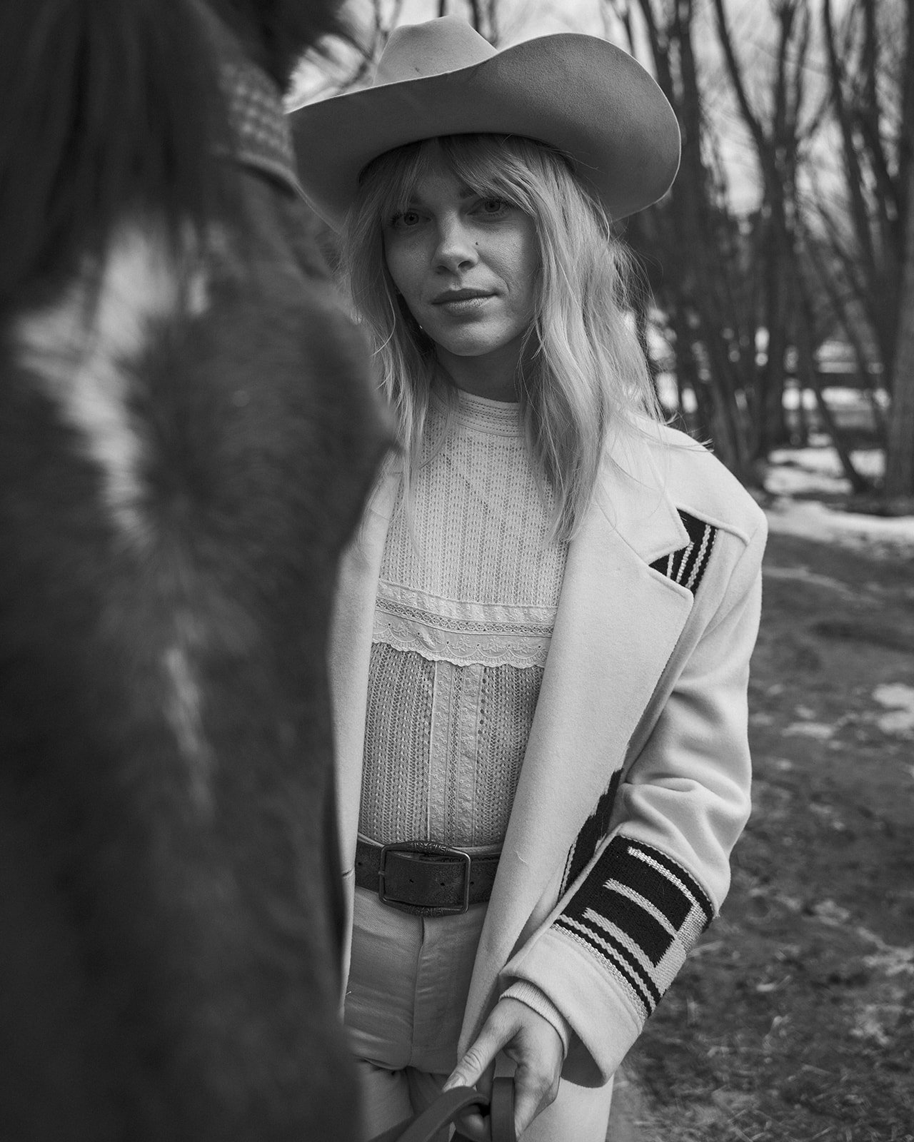 Black and white photo of a person wearing a cowboy hat and a patterned coat, standing outdoors with a horse in the foreground.