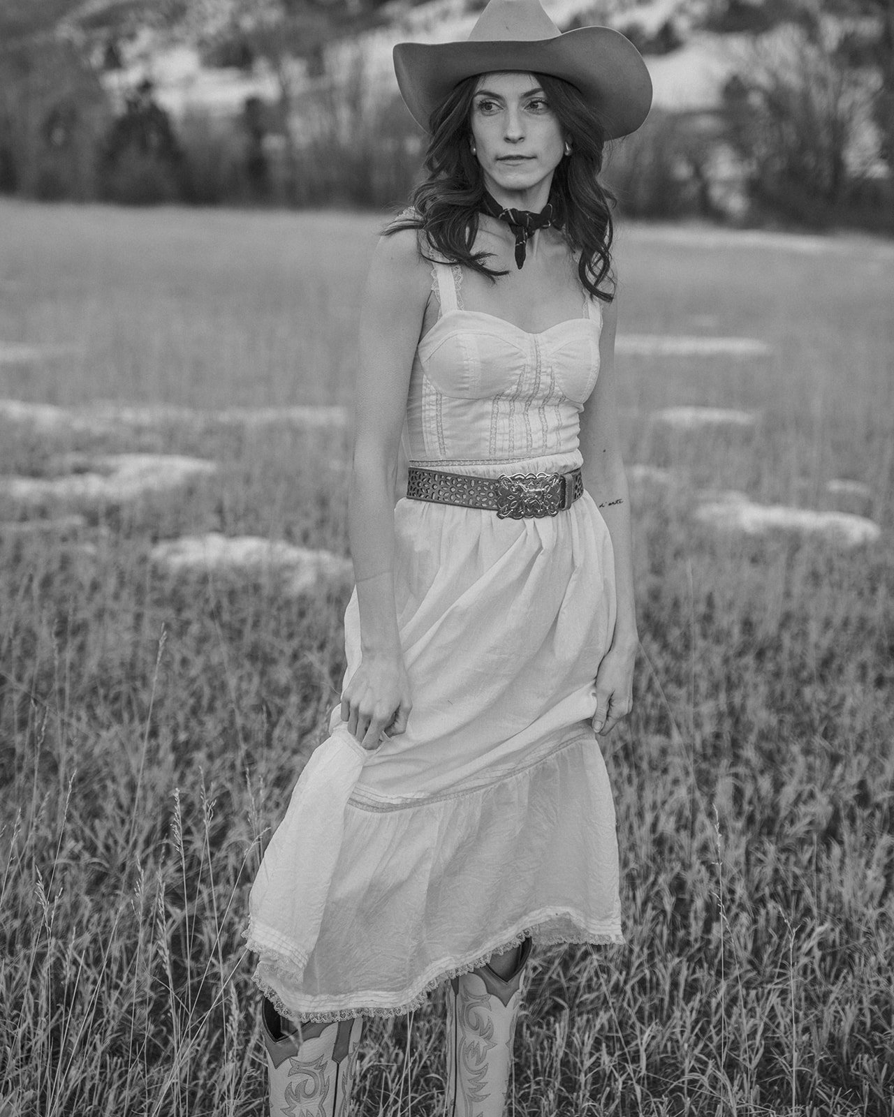 Woman in a field wearing a cowboy hat, dress, and cowboy boots, black and white photo.