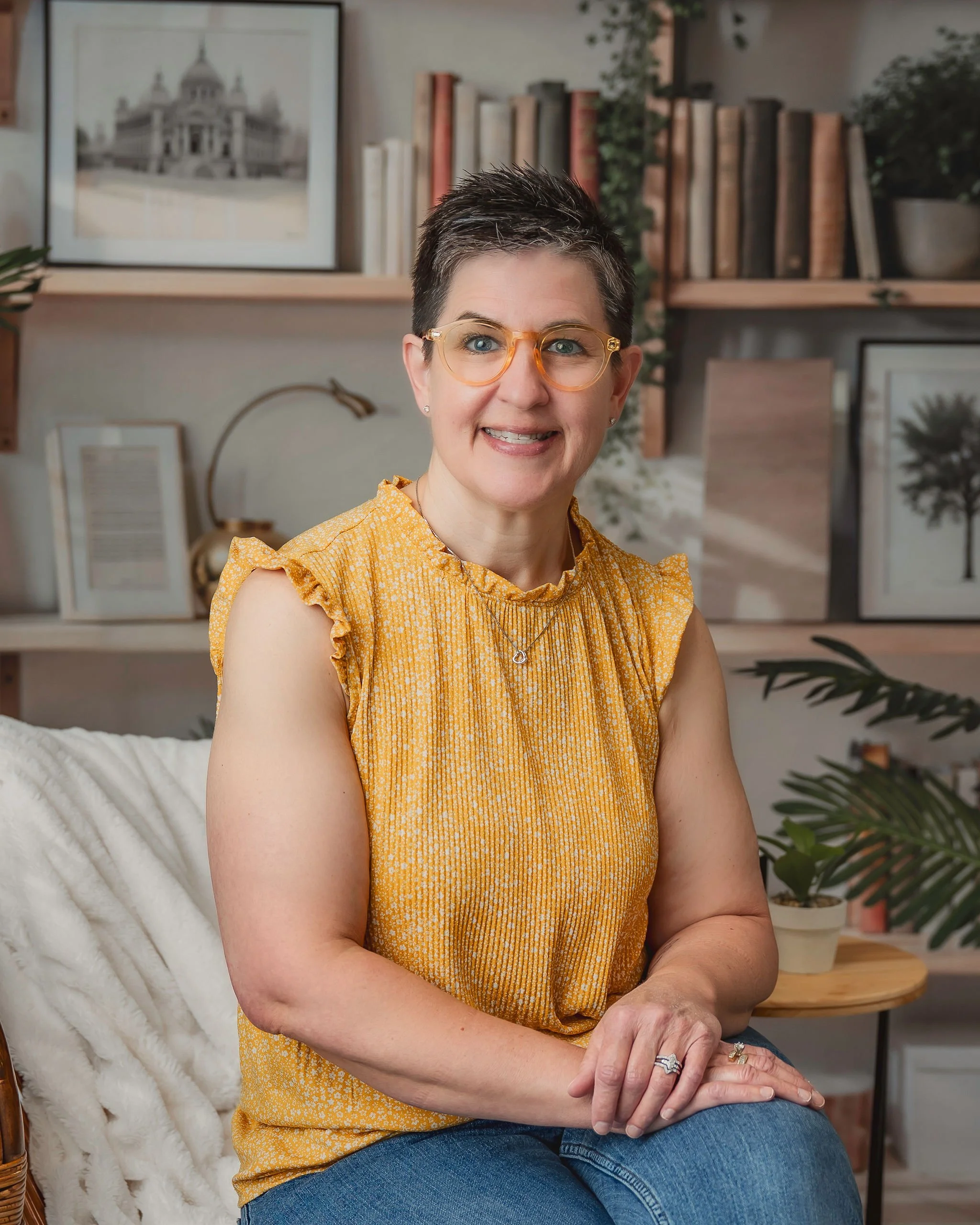 Smiling woman with short dark hair and glasses sitting on a cream-colored blanket in a cozy room with bookshelves, framed pictures, and plants in the background.