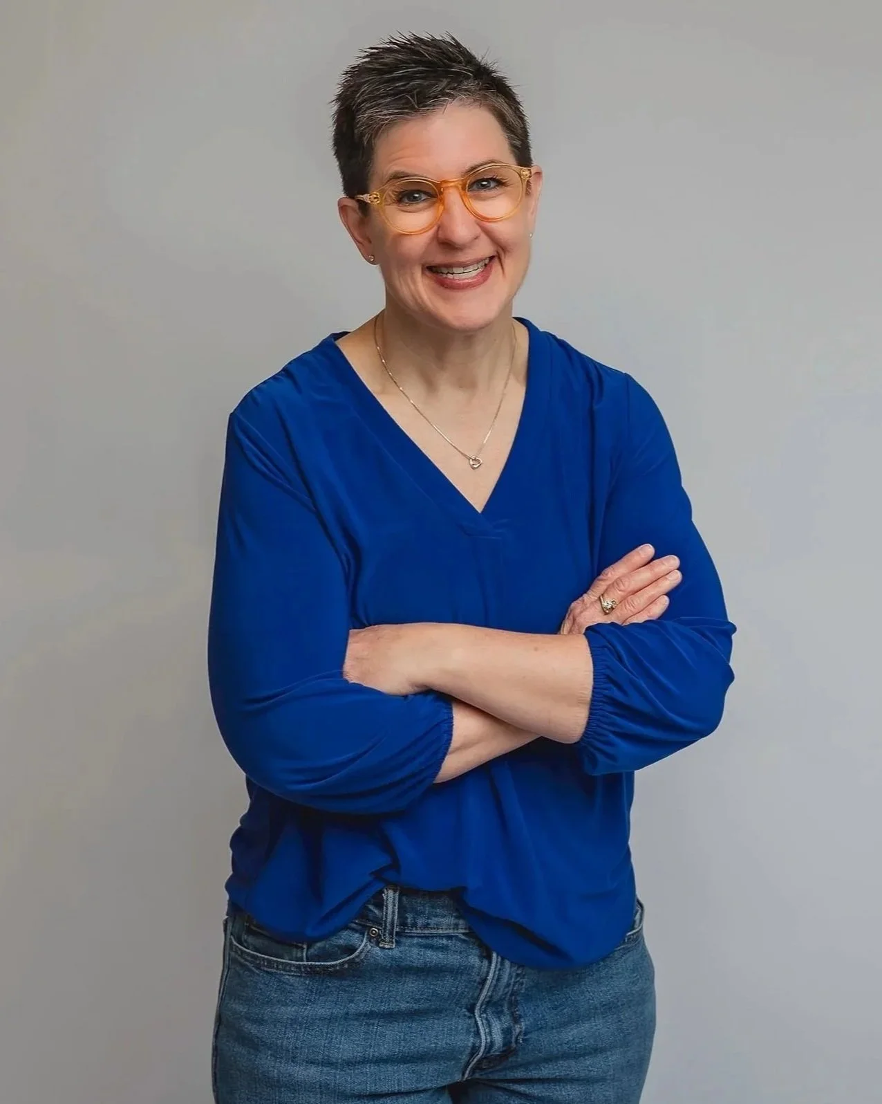 A smiling woman with short dark hair and glasses, wearing a blue blouse, crossing her arms, standing against a plain gray background.
