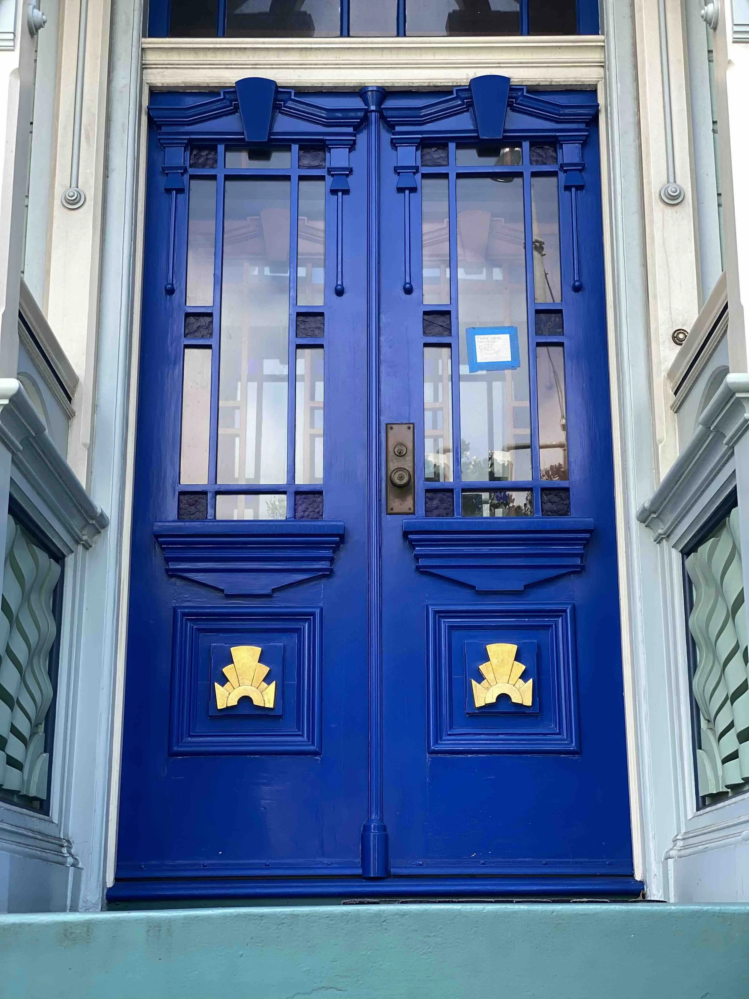 front door repair process on this striking royal bright blue Classic European double-door architecture with multi-pane glass upper sections and carved geometric wooden panels below