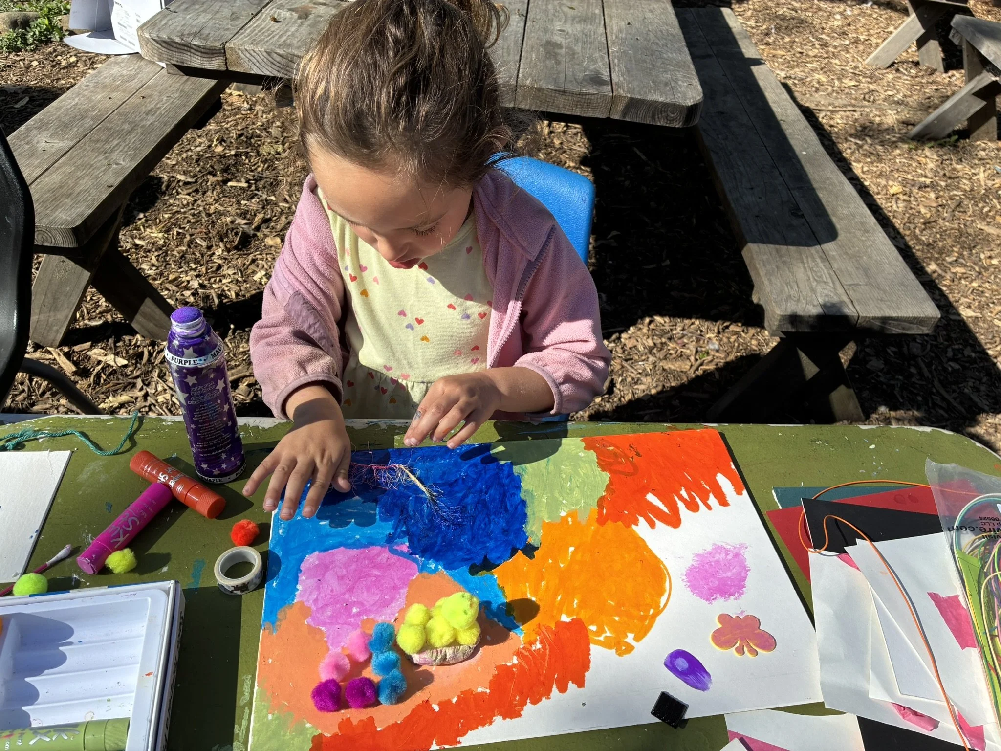 A young girl with brown hair is seated at an outdoor table, creating artwork with colorful paints. The table is covered with various art supplies, including pom-poms, markers, paint, and paper. Behind her is a wooden picnic table and one of its attached benches, set on a wood chip ground with shadows cast by nearby trees.